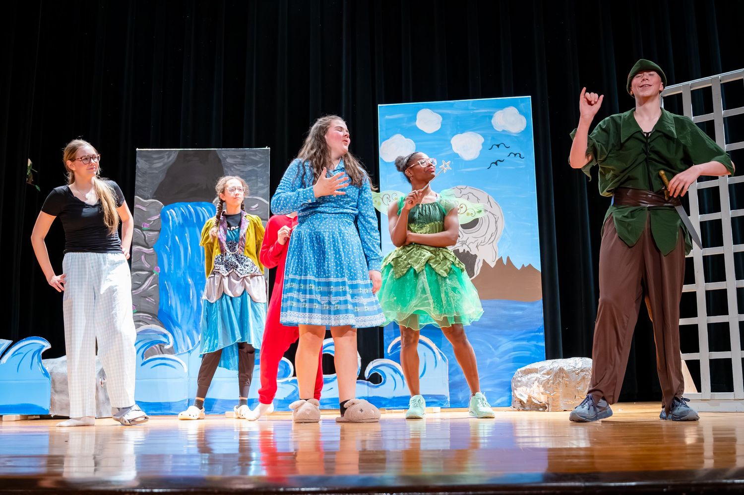 Performers in colorful costumes act on a stage with a blue backdrop and white set pieces during a theatrical production.