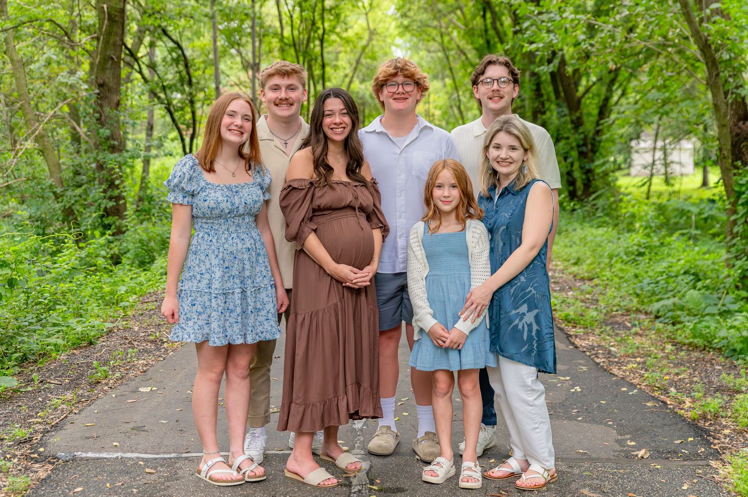 A group of people wearing coordinated blue and brown outfits pose together on a tree-lined path during a photo session.