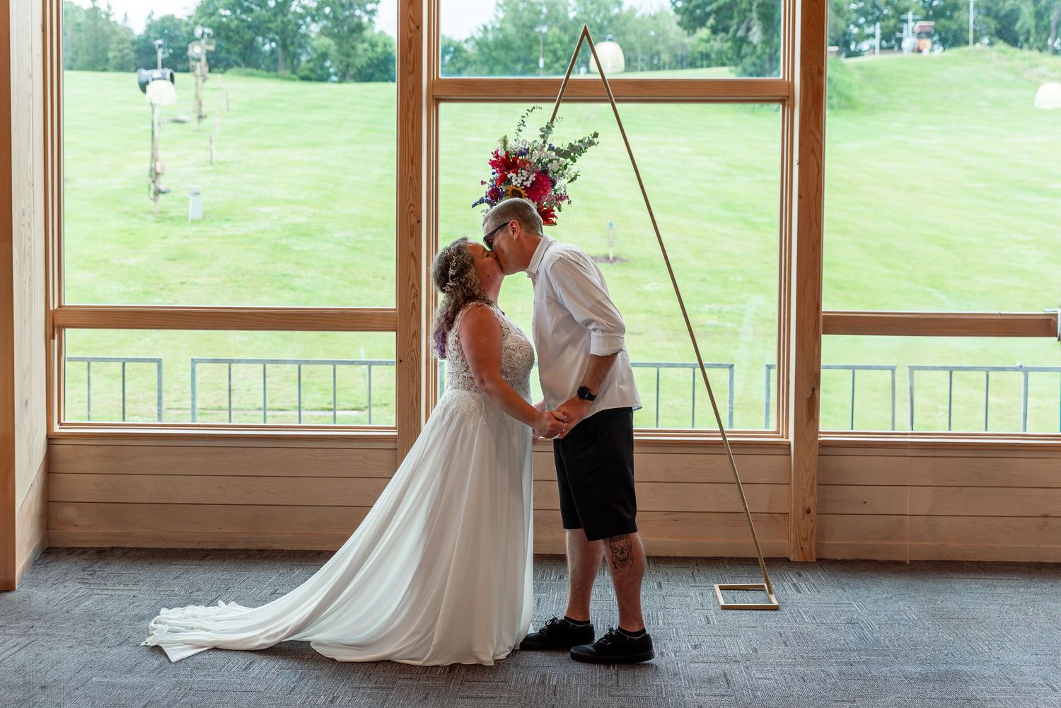 A wedding couple shares an intimate moment in a rustic barn setting with large windows overlooking green pastures.