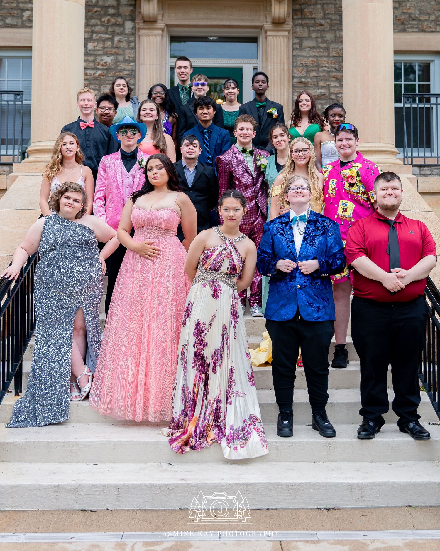 A group in formal attire poses together on stone steps outside a brick building for a special occasion.