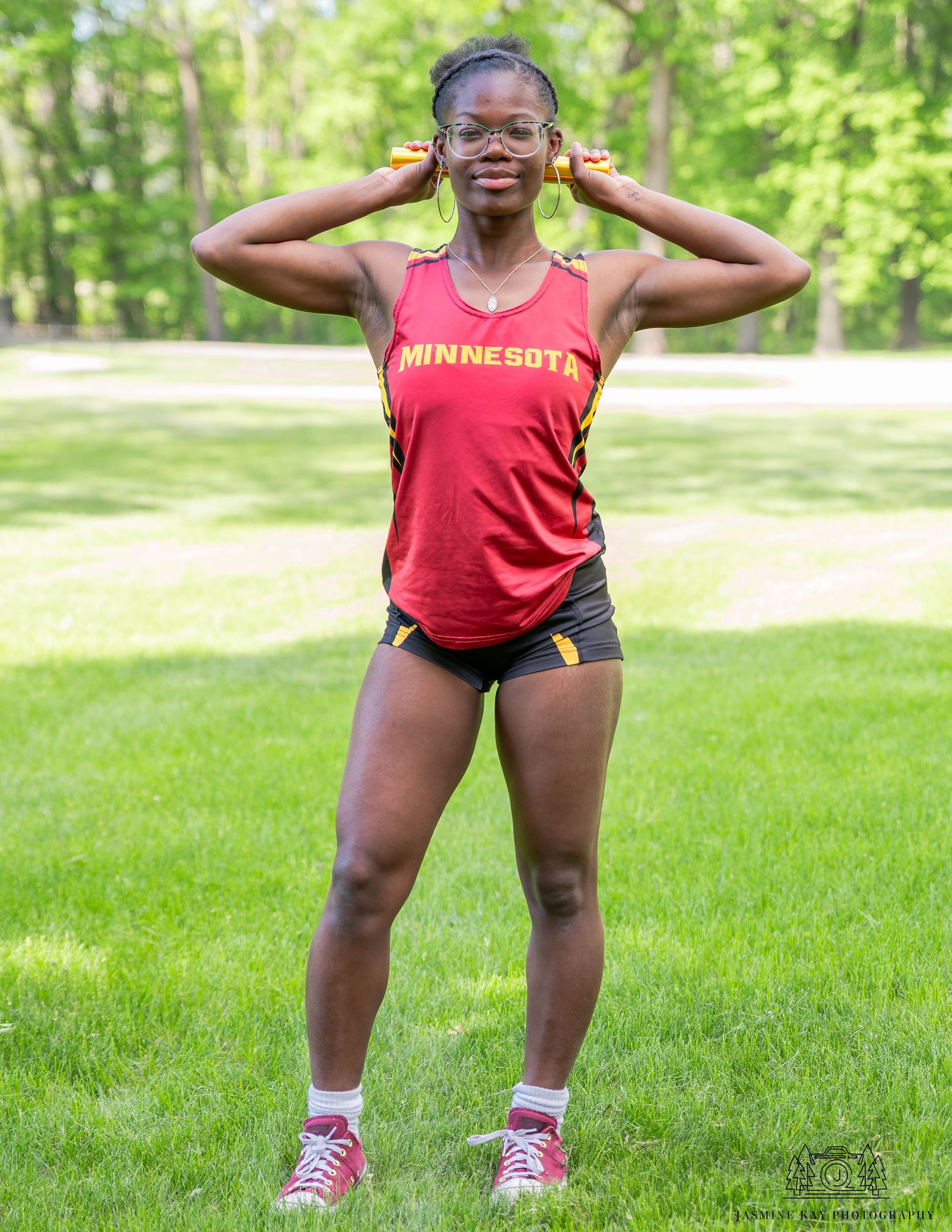 Athlete in red Minnesota tank top and black shorts poses outdoors on grass with hands by ears.