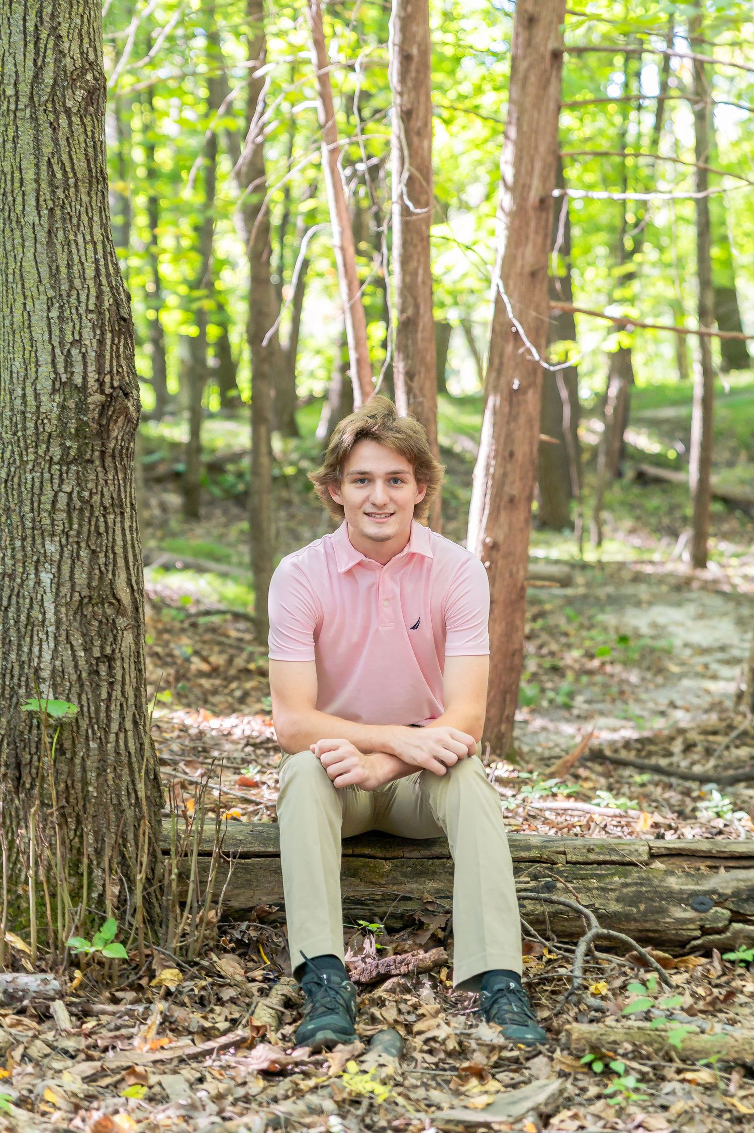 Person in pink shirt and khaki pants sits on fallen log in sunlit forest setting.