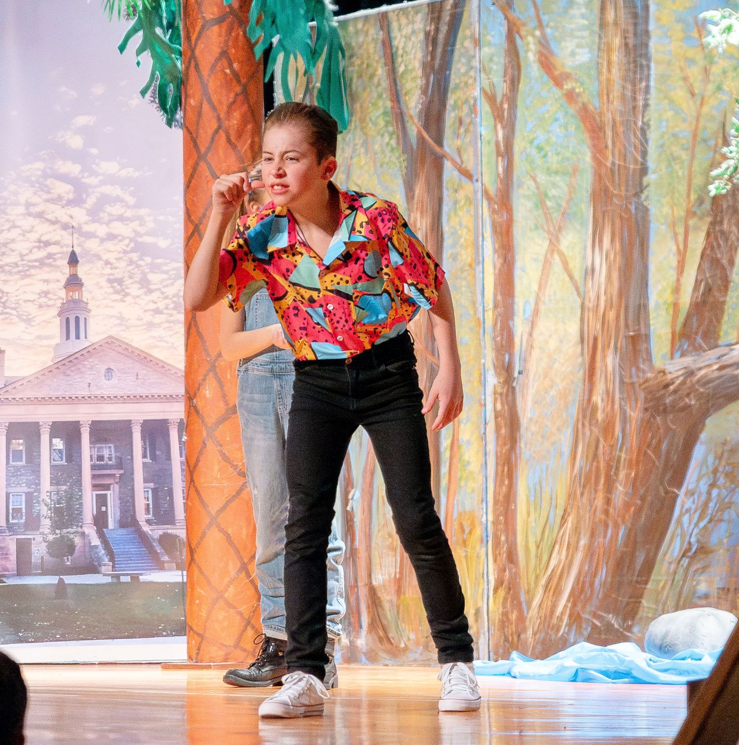 Performers dancing on a theater stage with colorful scenery and trees in the background.