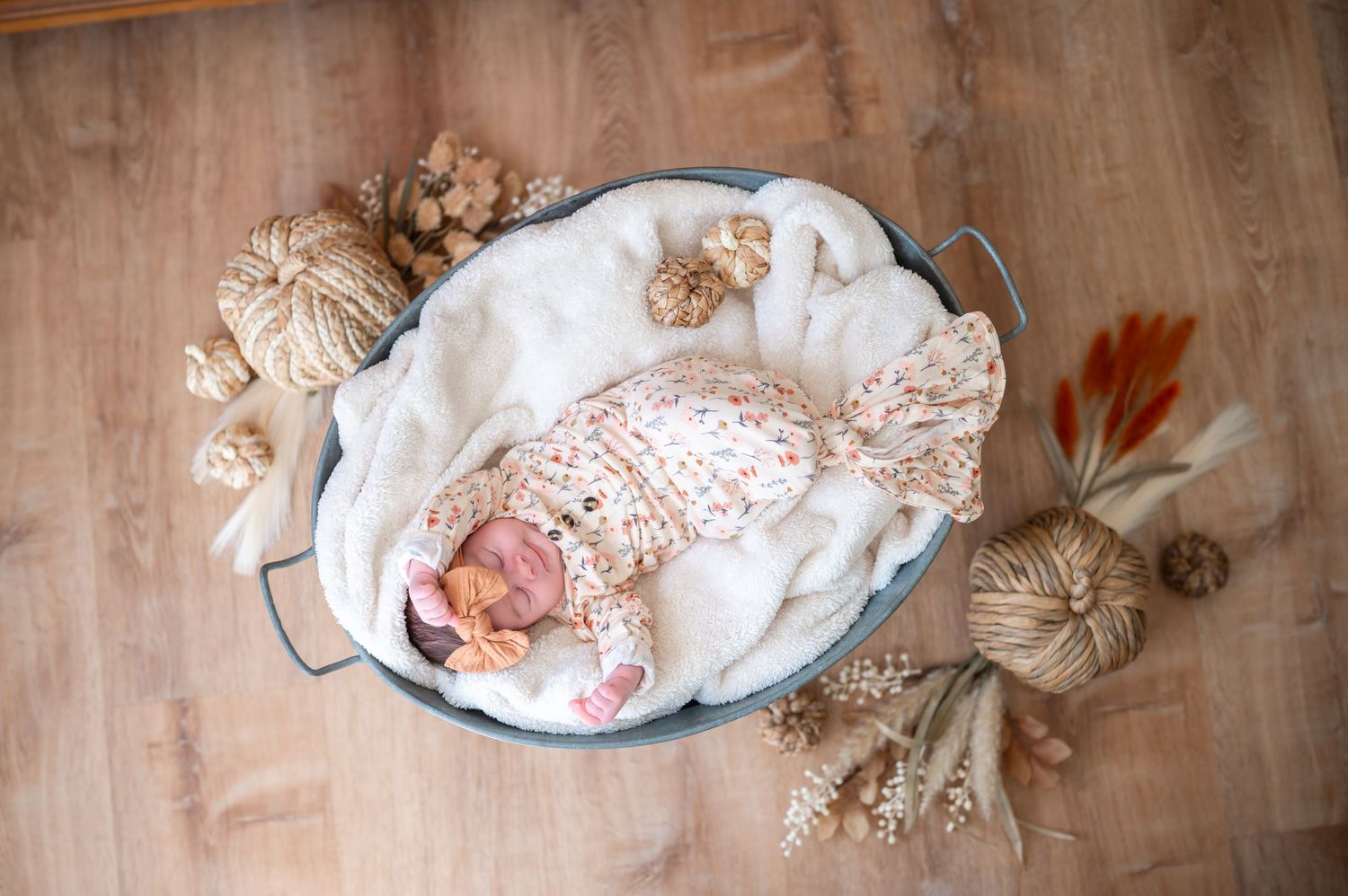 Sleeping newborn in white lace outfit lying in vintage enamel basin surrounded by dried flowers.