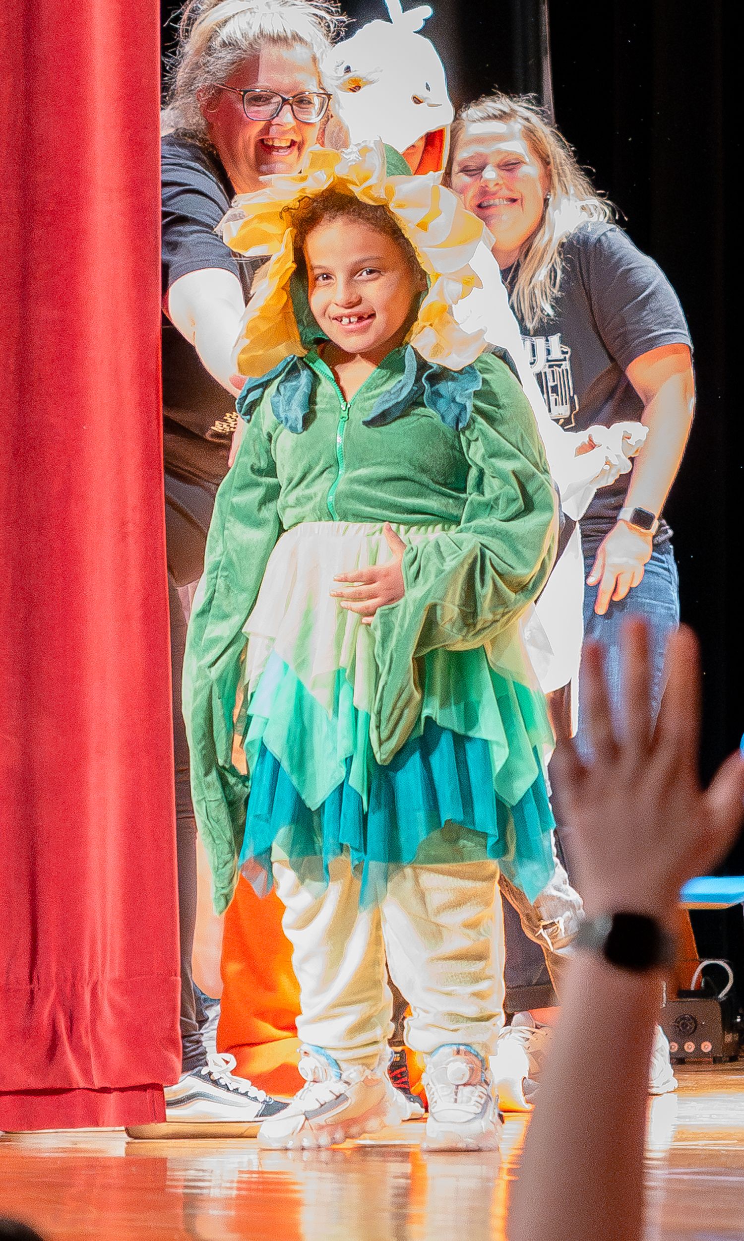 Children performing on stage wearing green costumes with red curtains in the background.
