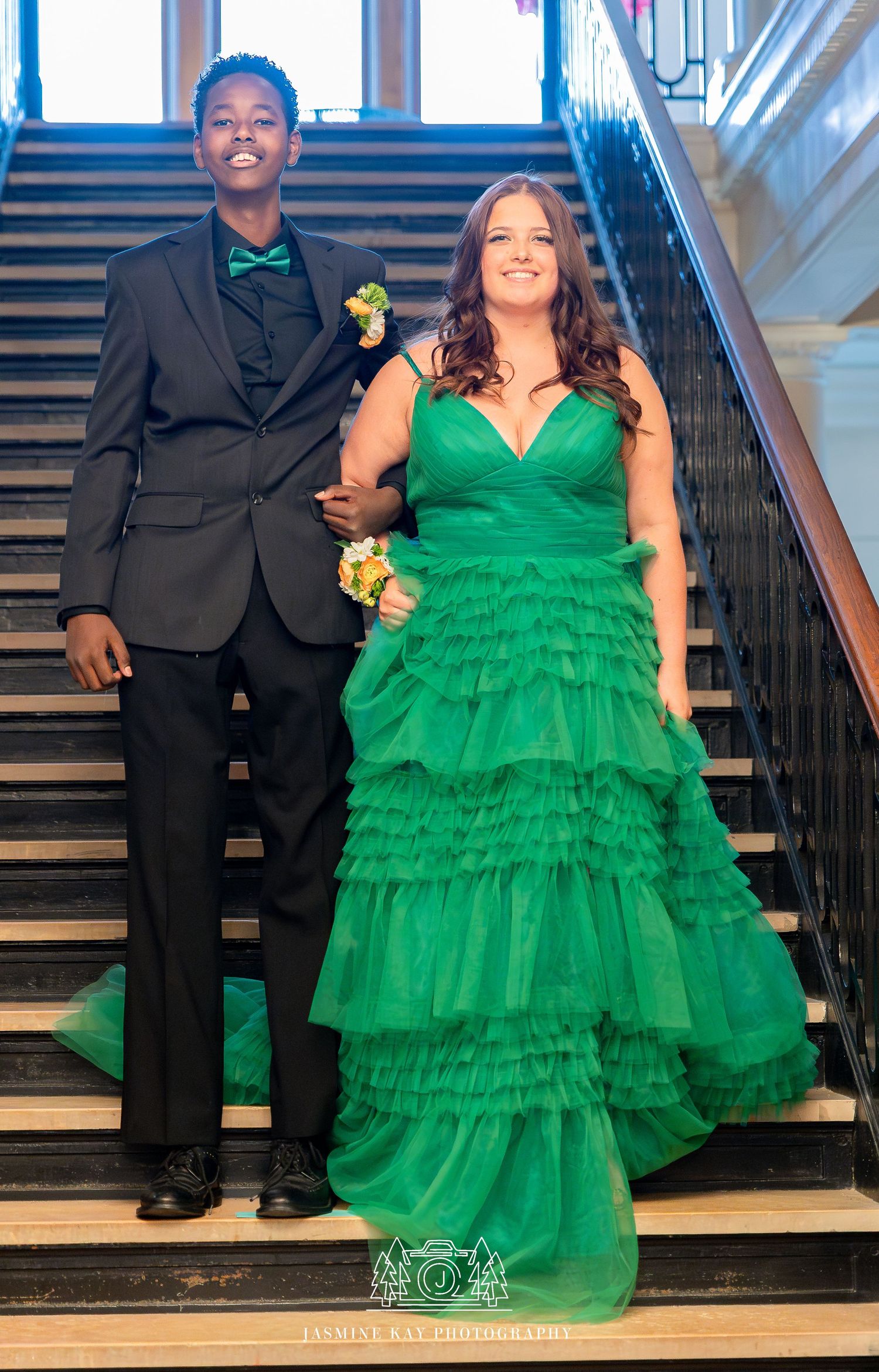 Two people in formal attire stand on elegant stairs, with one wearing a ruffled emerald green gown and yellow corsage.