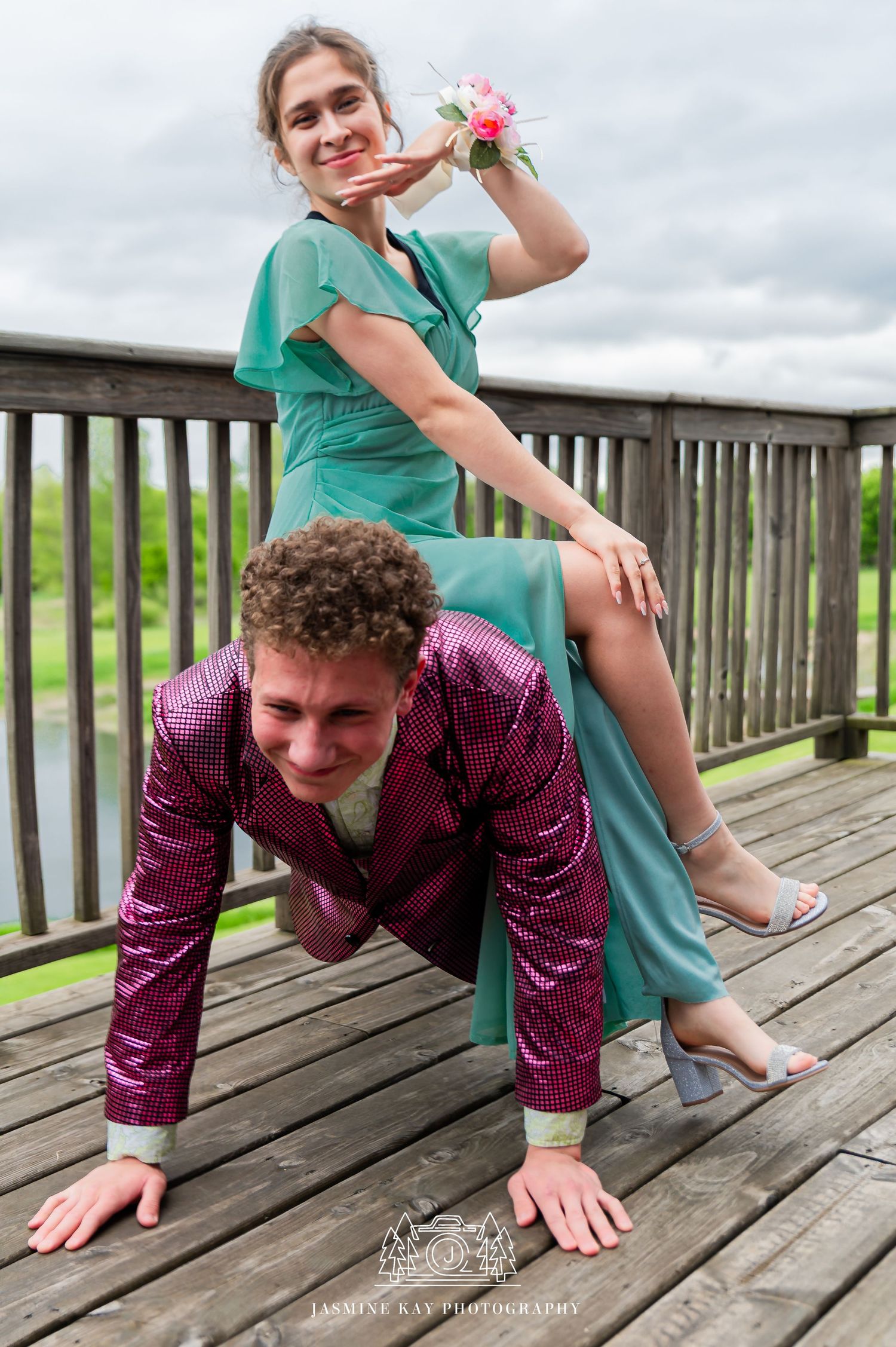 Two people in formal attire pose playfully on a wooden deck overlooking a cloudy landscape.