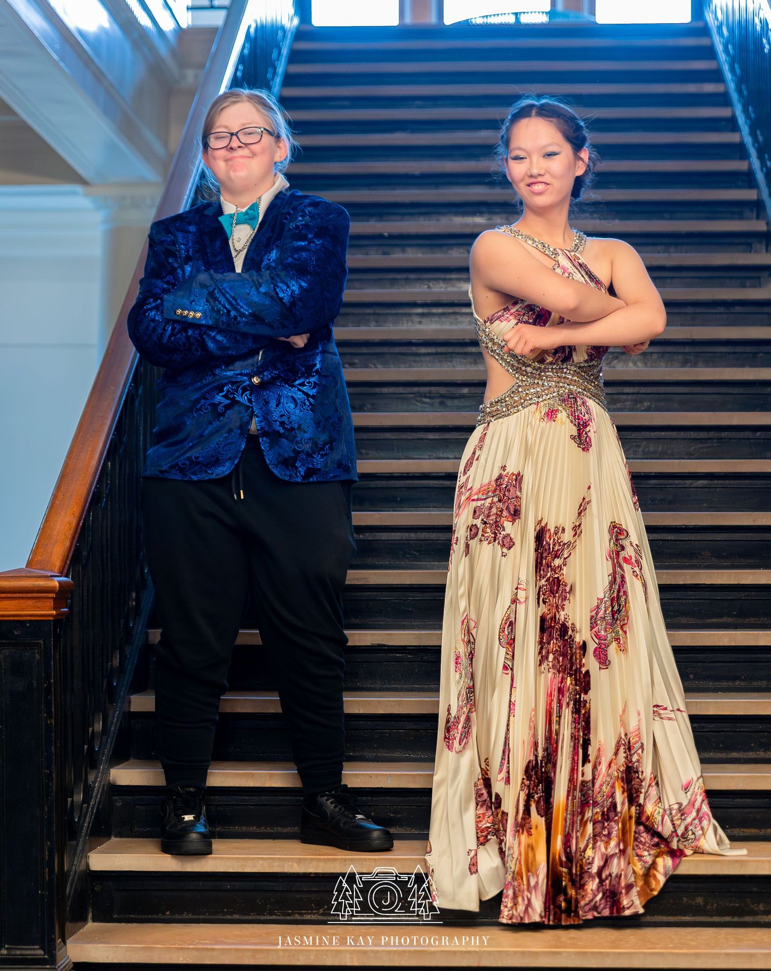 A couple sits together on wooden stairs wearing formal attire including a floral dress and dark suit.