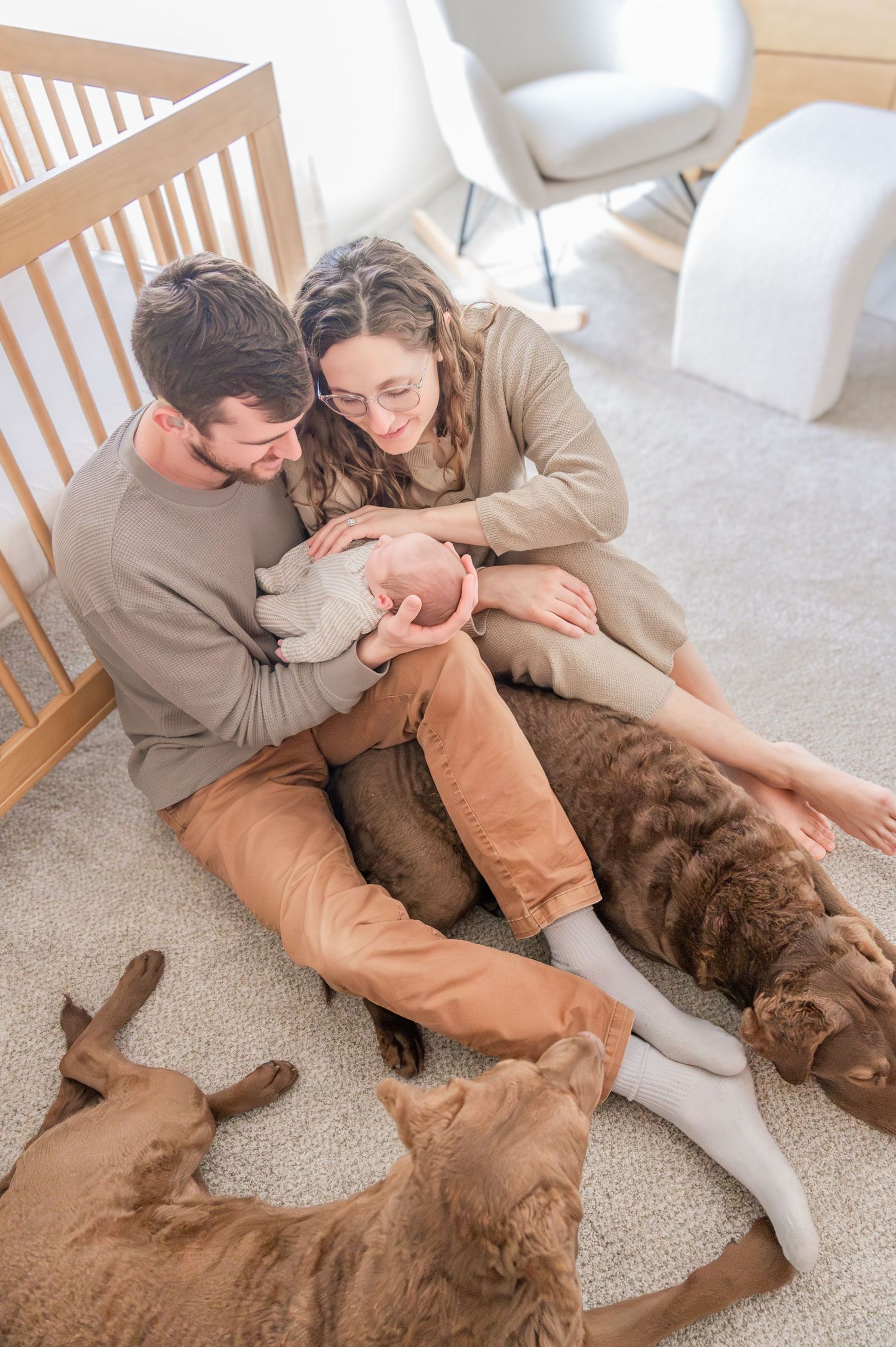 A couple sits on carpet with their sleeping baby while two brown dogs rest peacefully beside them.