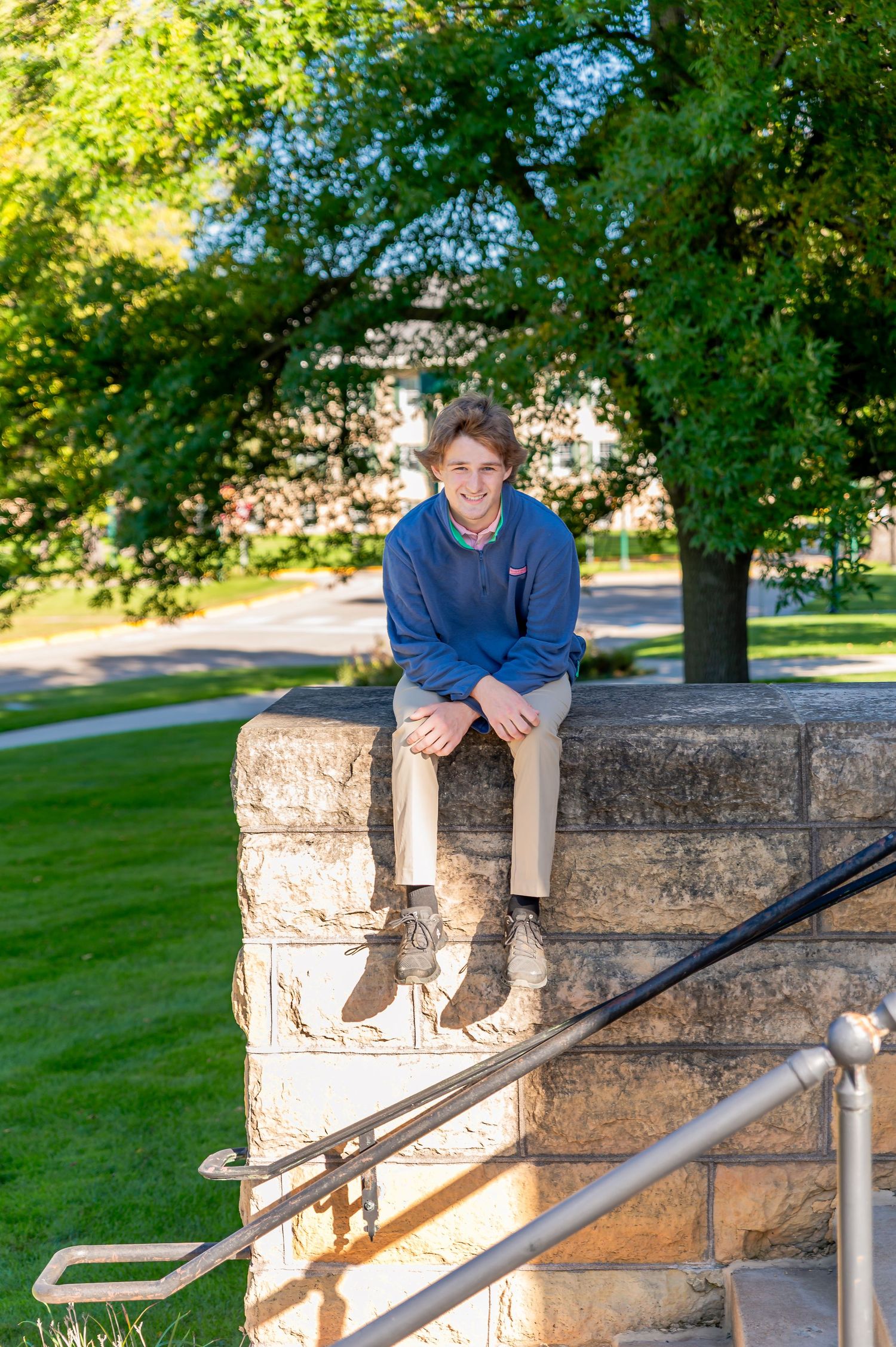 A person in a blue shirt sits casually on a stone wall with metal railings in a sunny park setting.