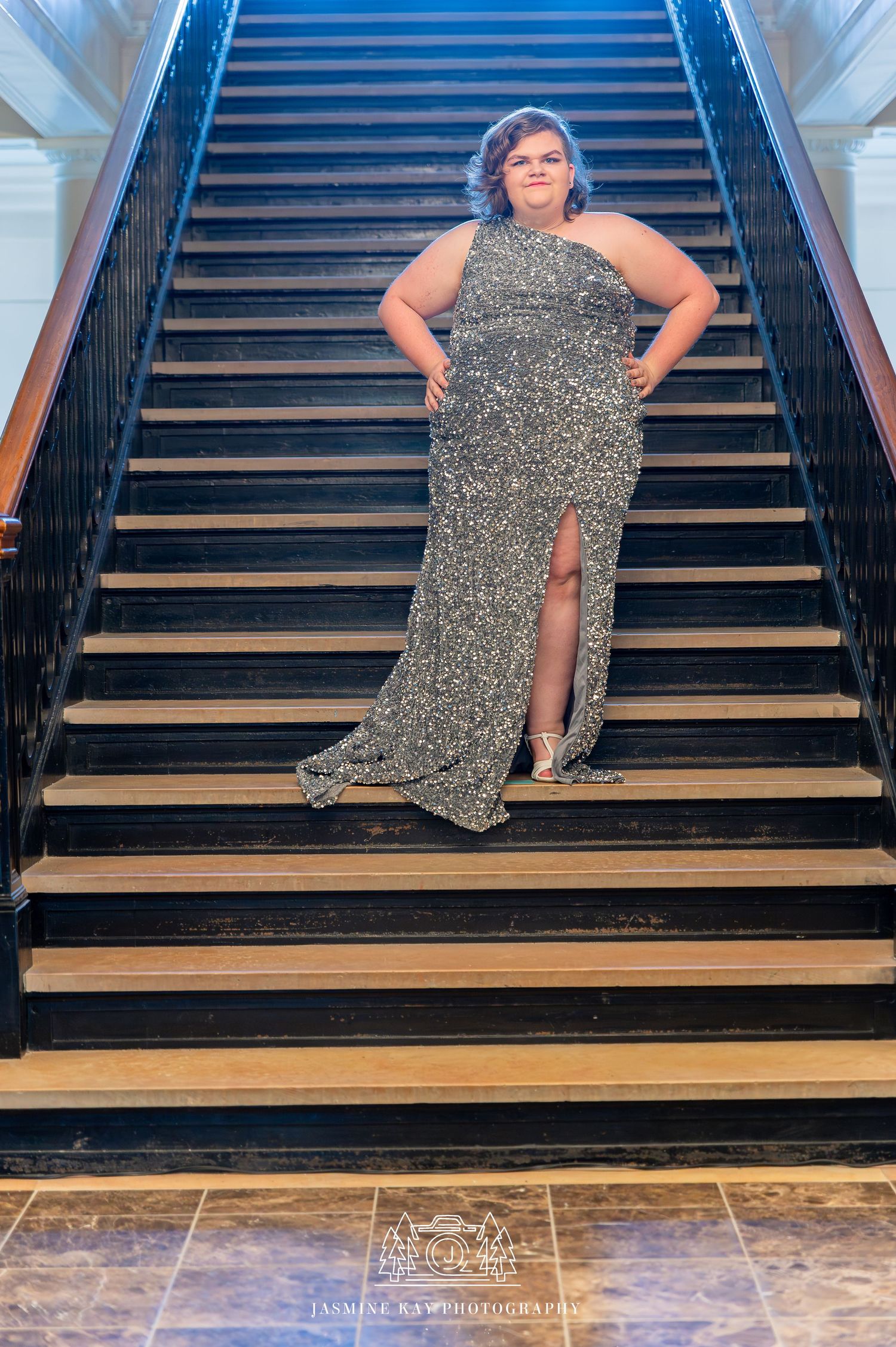 A person in a sparkly silver evening gown with a thigh slit poses confidently on an elegant staircase.