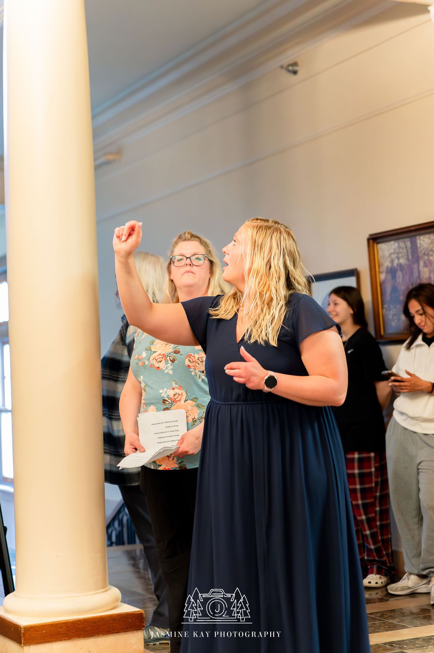 A woman in a navy blue dress gestures animatedly while speaking in an indoor venue with columns and framed artwork.