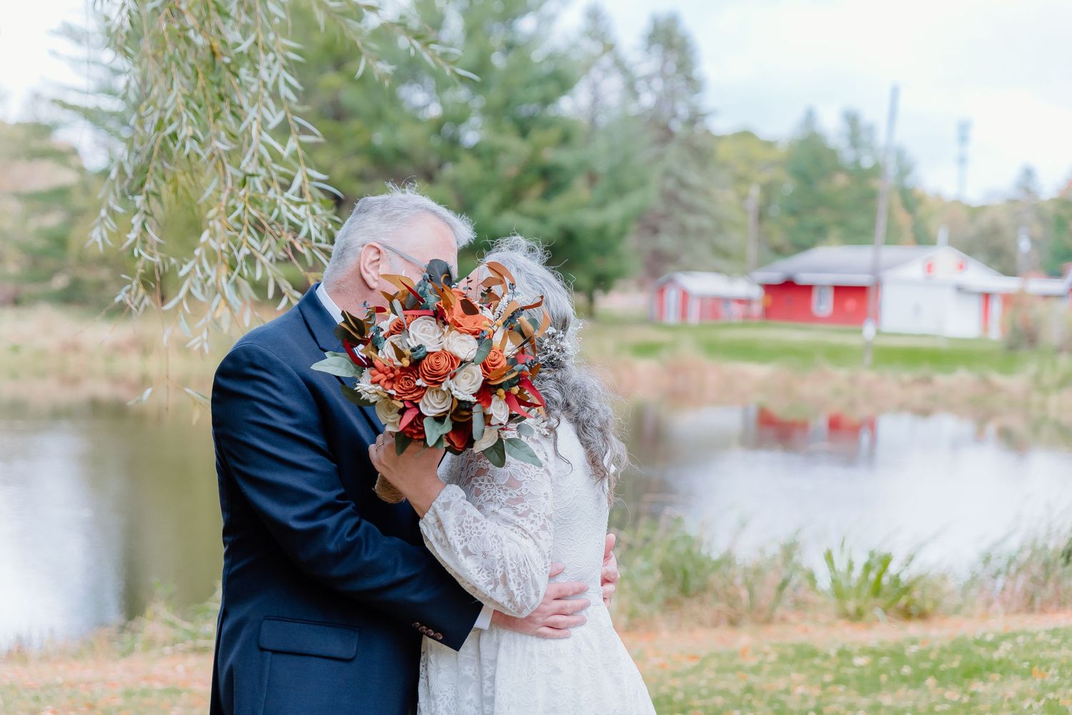 Romantic autumn wedding portrait by a pond with red and orange bouquet.