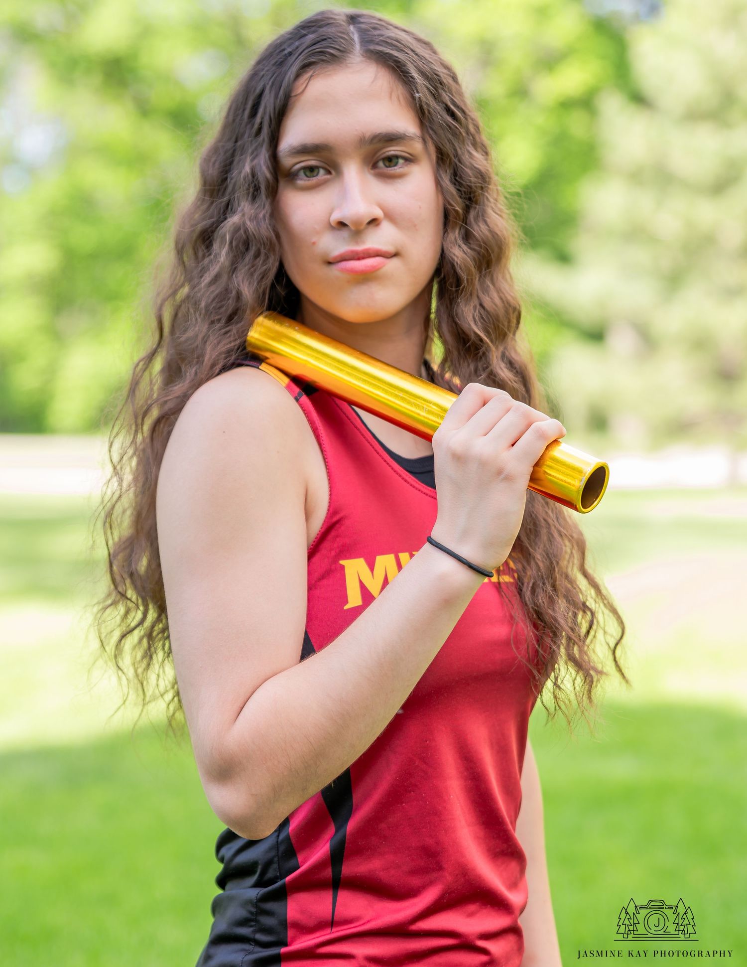 Person wearing red and yellow athletic uniform poses outdoors in a park setting with green foliage in background.