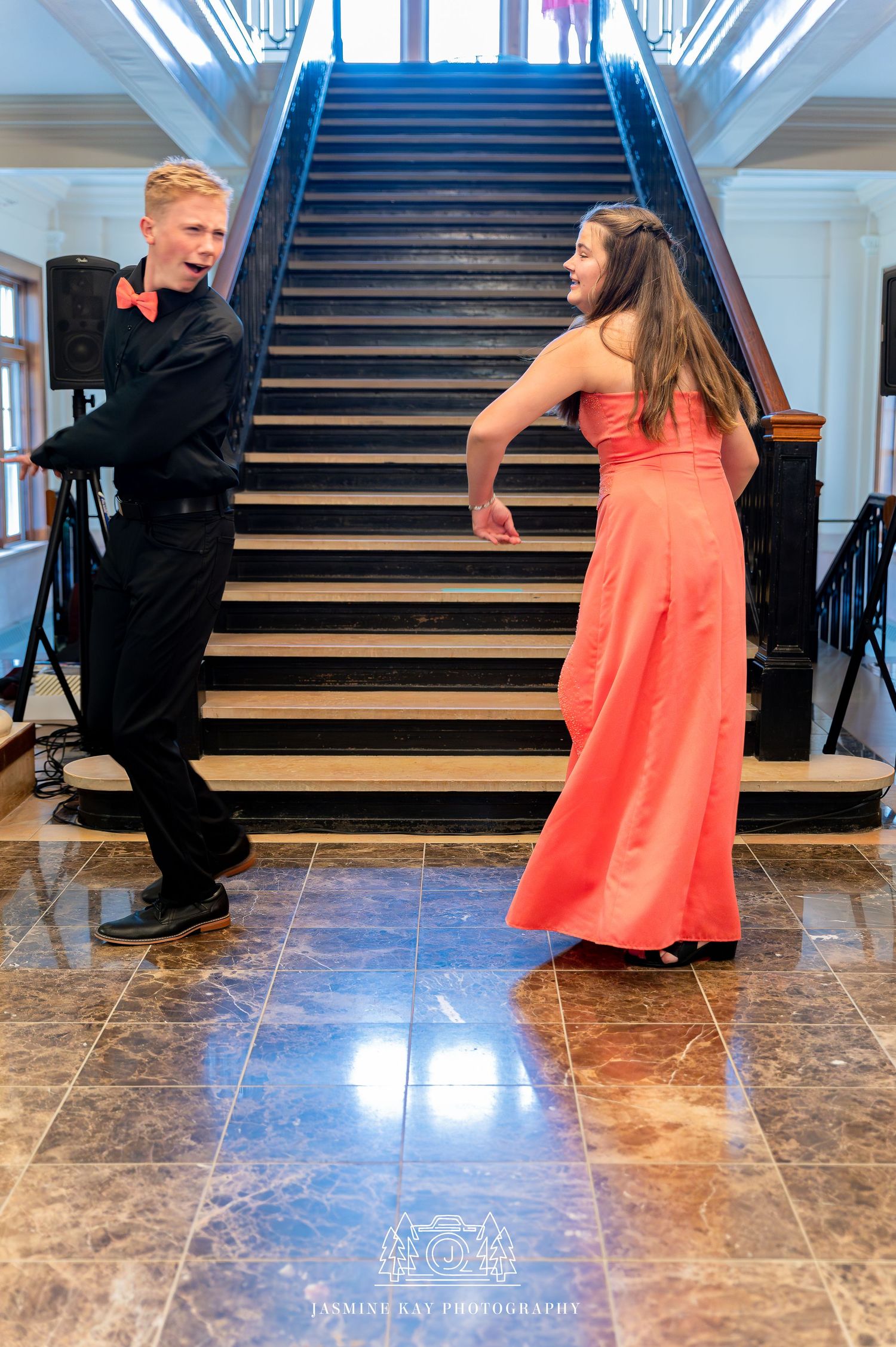 A person in an orange dress dances with someone in black formal wear on marble floors near elegant stairs.