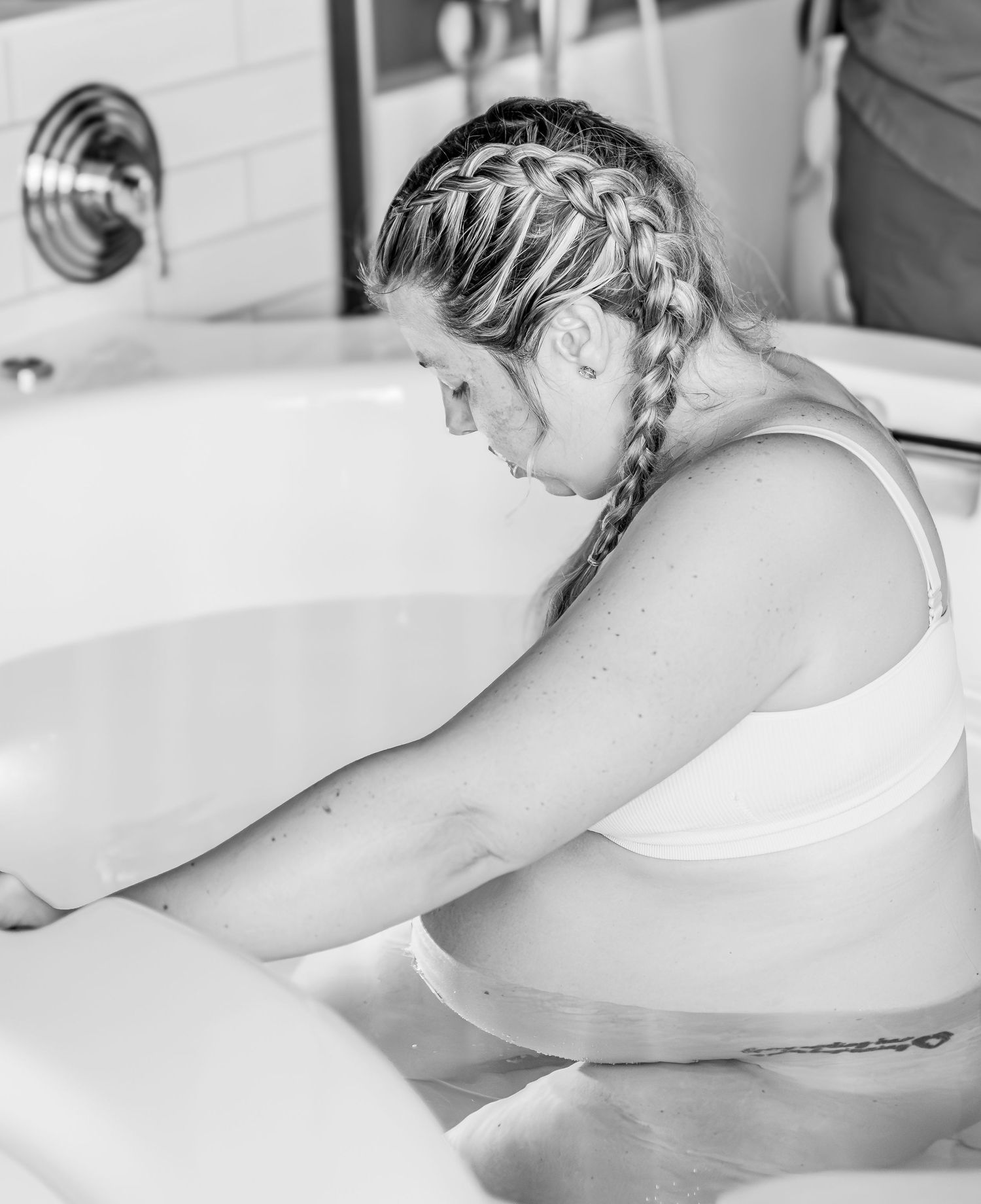 Black and white photo showing someone with braided hair in a white tank top leaning over a bathtub.