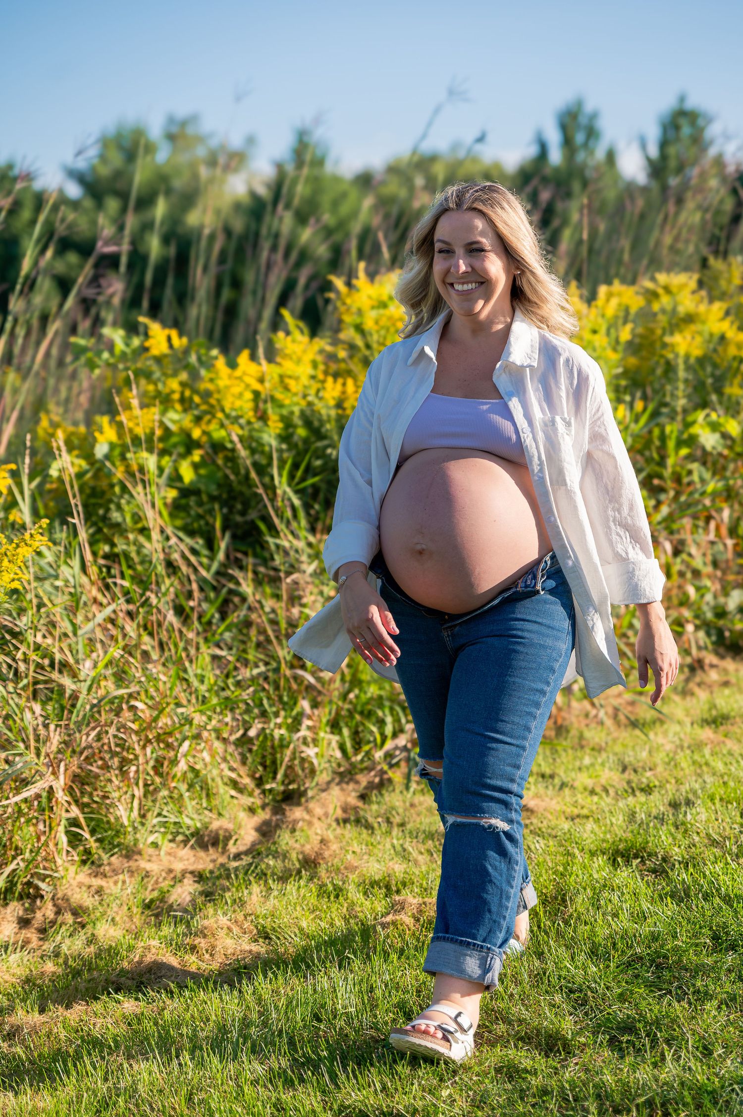 Expectant mother walks through a sunny meadow with blooming yellow wildflowers on a bright summer day.