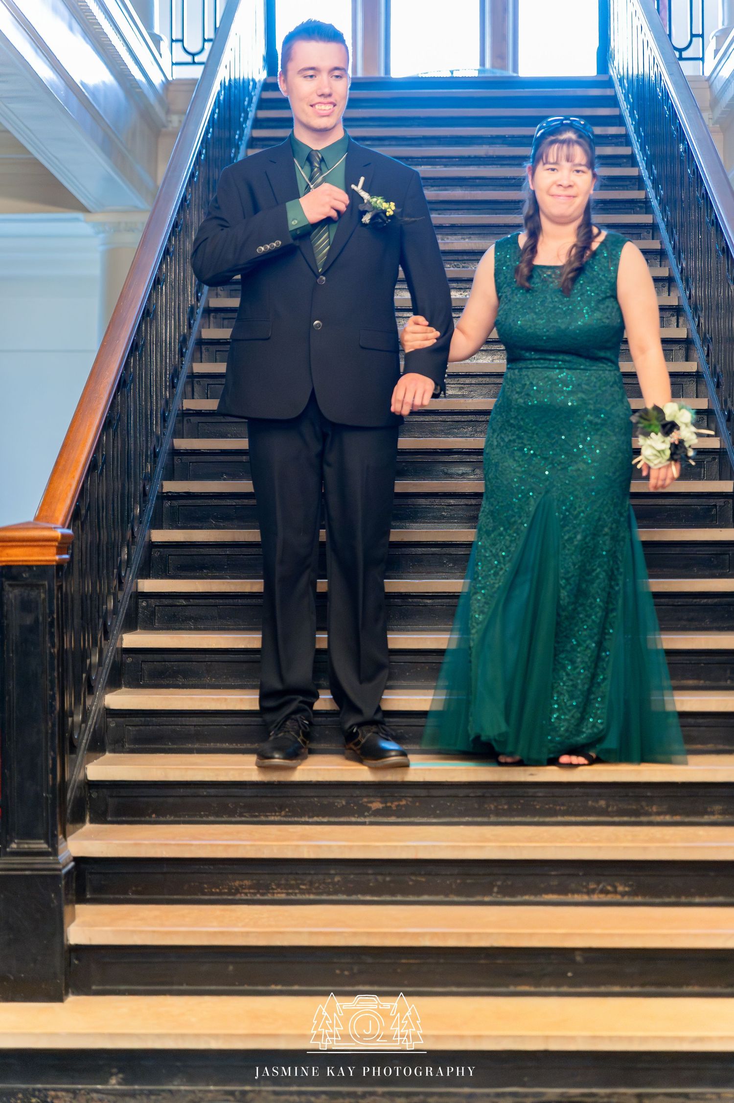 A couple in formal attire stands on a grand staircase, with the woman wearing a sparkly emerald green gown.