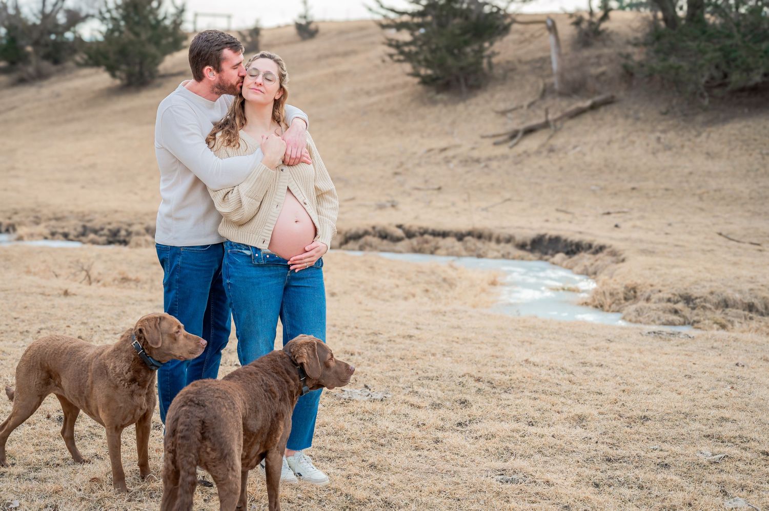 A couple poses outdoors with two Chesapeake Bay Retriever dogs in a winter landscape.