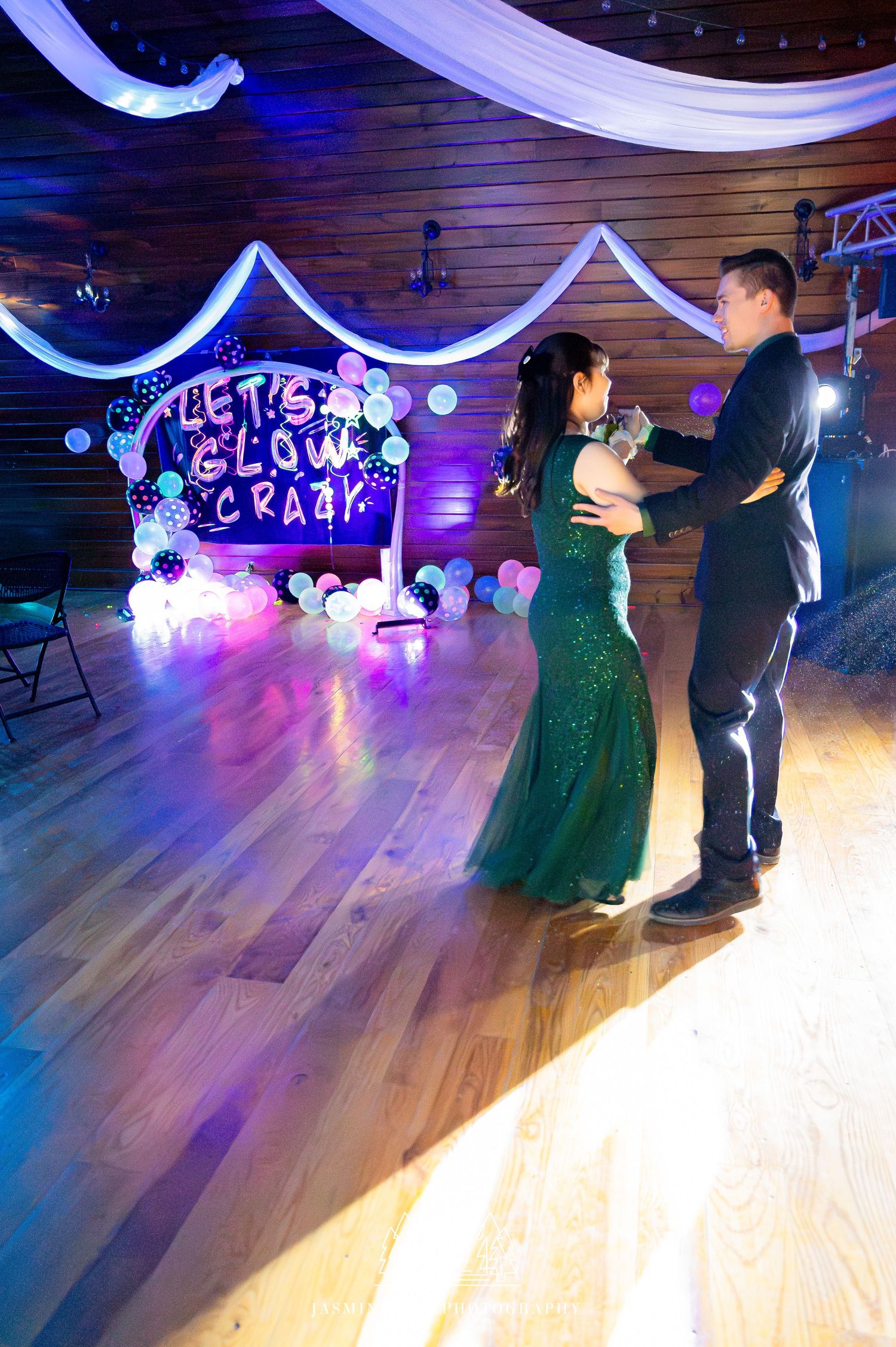 Couple dancing on a wooden floor under blue lighting with decorative white drapes and illuminated signage in the background.