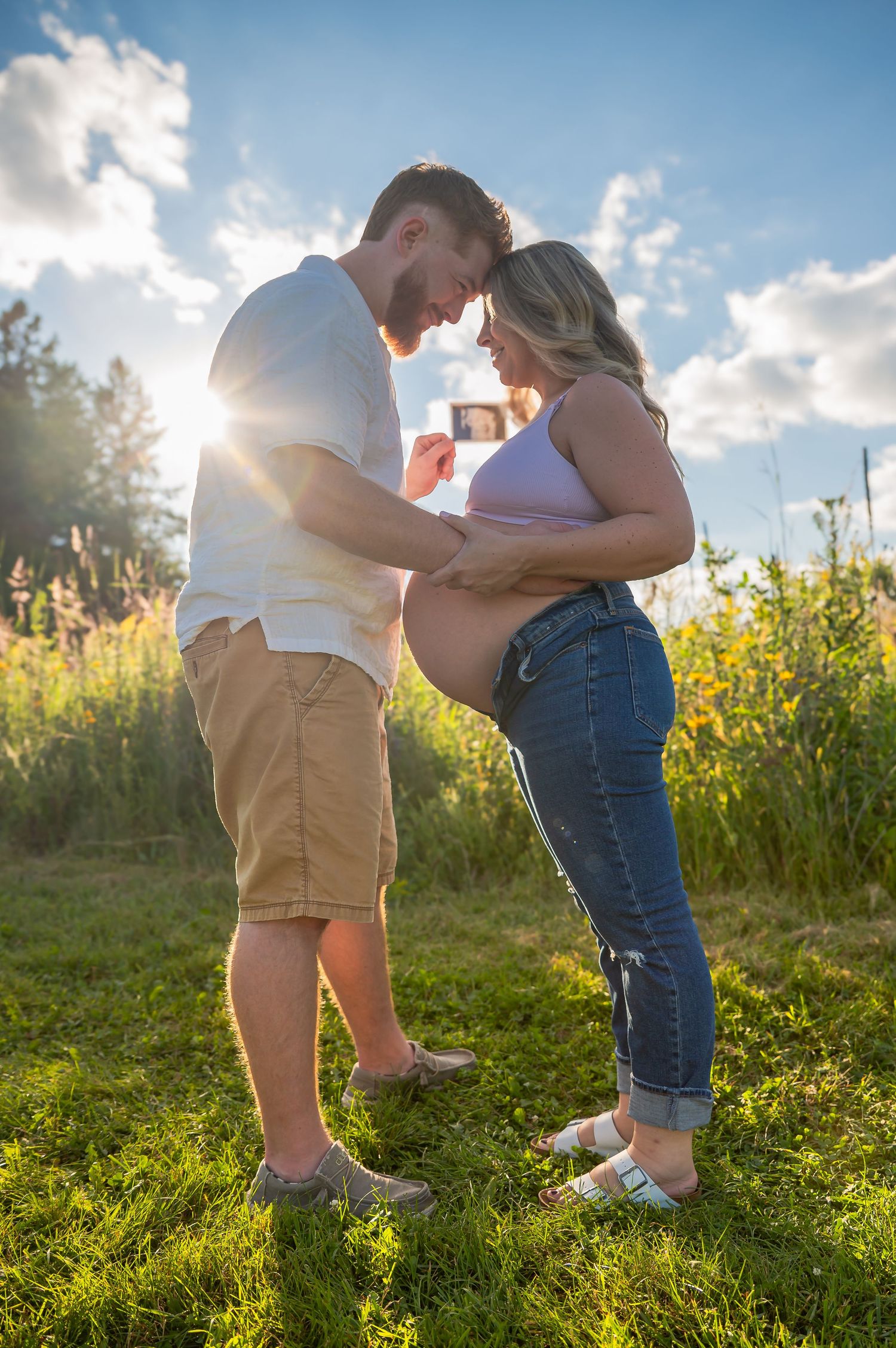 A couple embraces in a sunlit meadow with wildflowers during golden hour, creating a romantic outdoor scene.