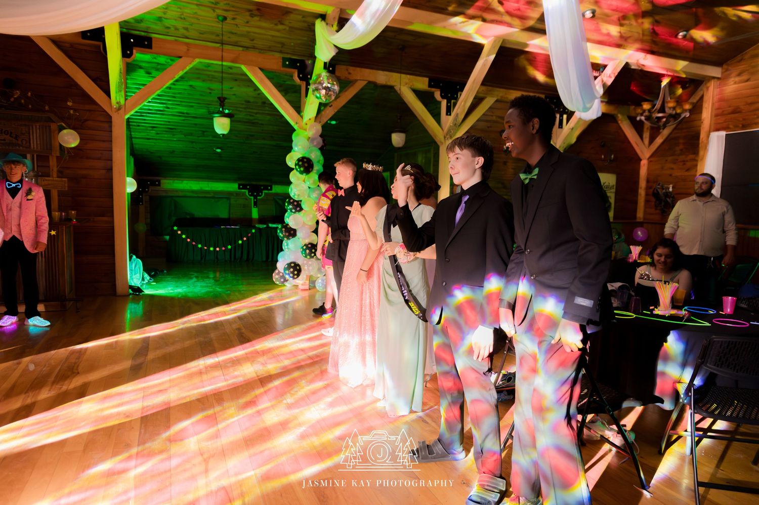 Dancers move across a rustic wooden dance floor illuminated by colorful lights at a wedding reception.