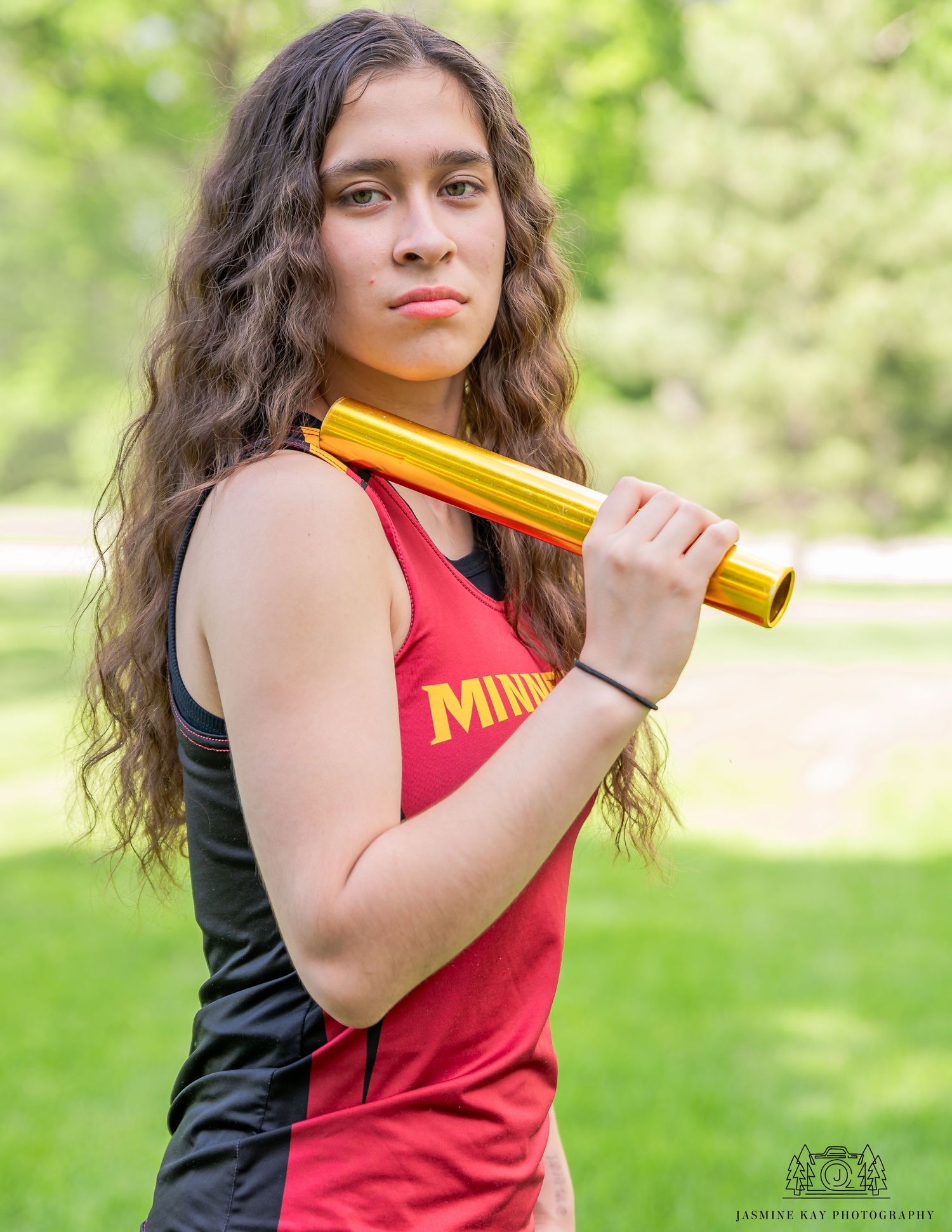 A person in a red sleeveless top practices softball batting stance outdoors in a grassy area during summer.