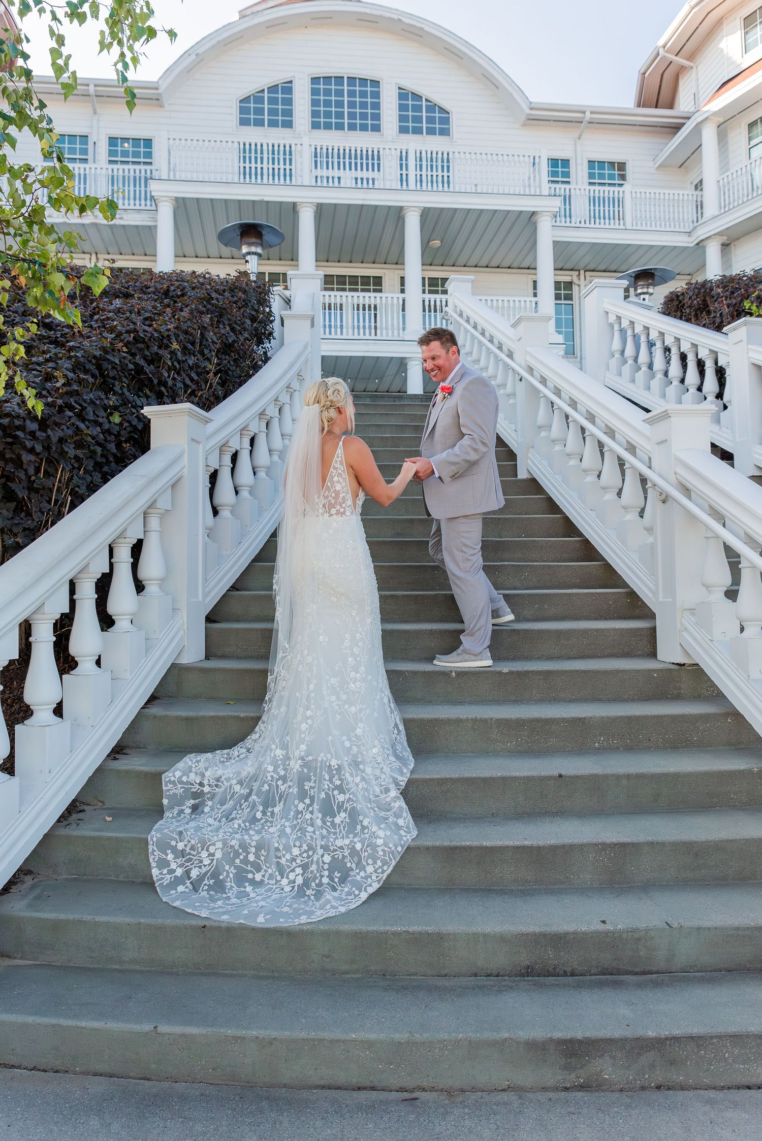 Bride in lace gown and groom on elegant white stairs leading to classic building.