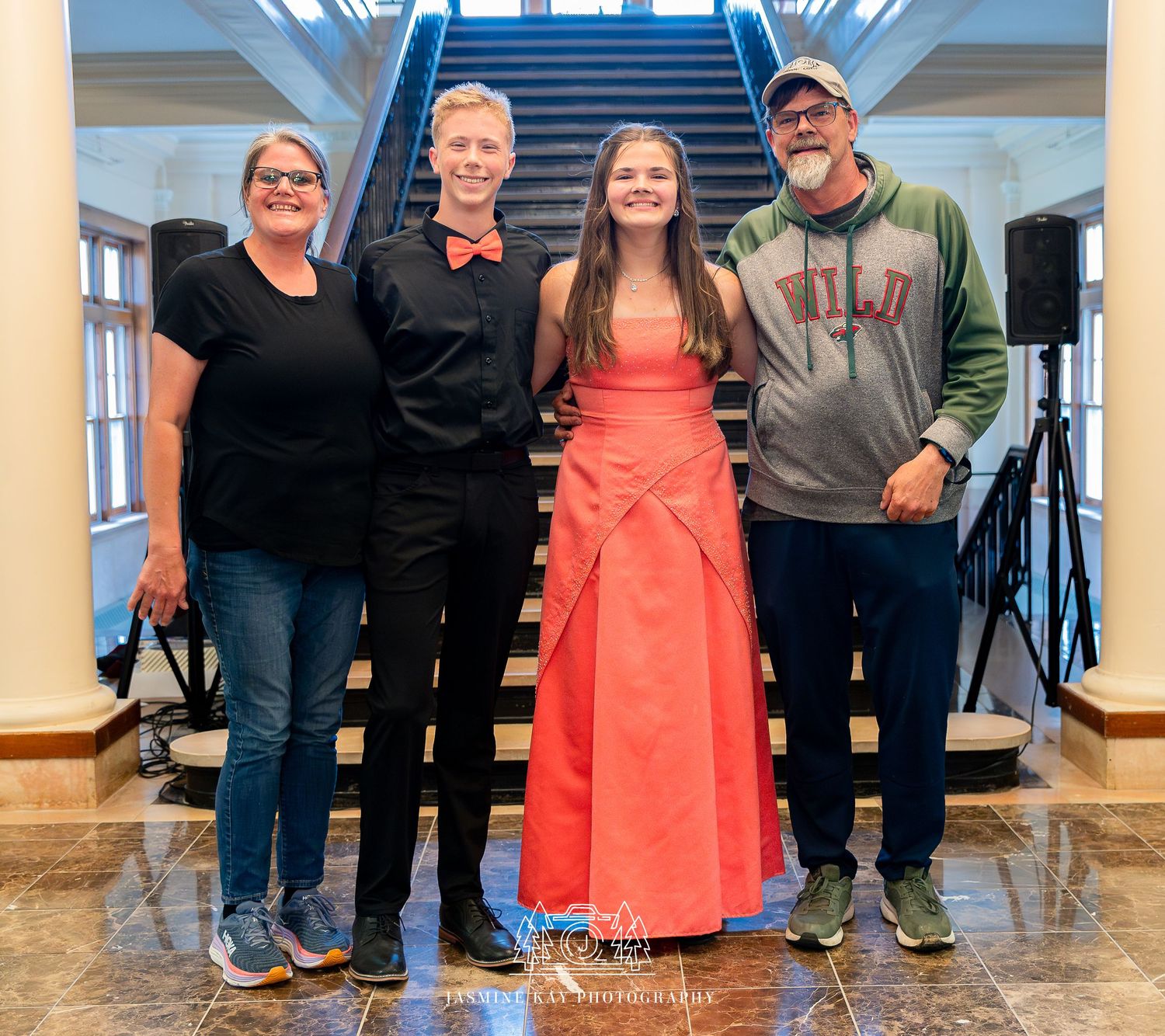 A group of four people stand together at the bottom of a staircase, with one wearing a bright coral formal dress.