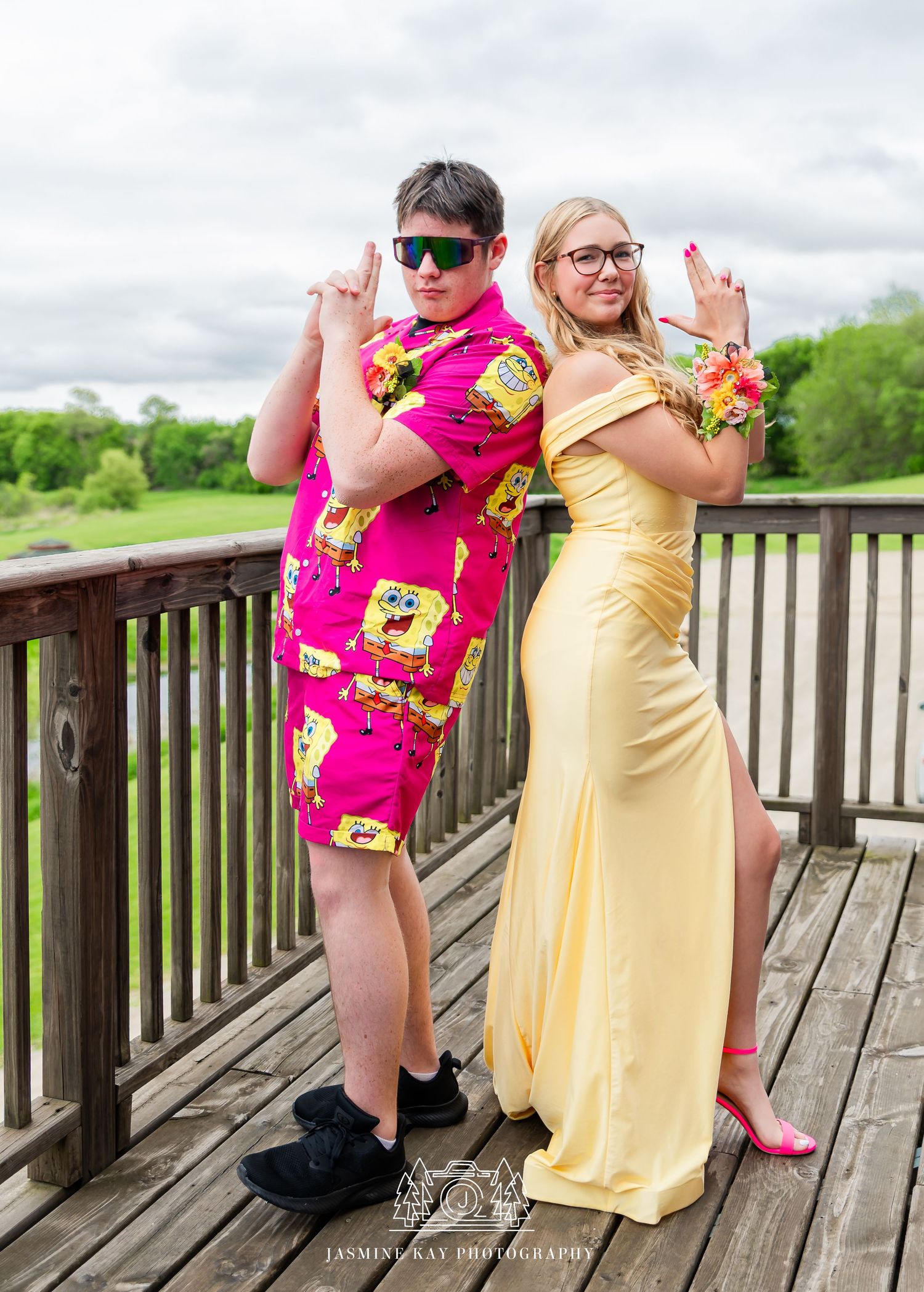 Two people in bright pink and yellow dresses strike playful poses on a wooden deck overlooking green scenery.