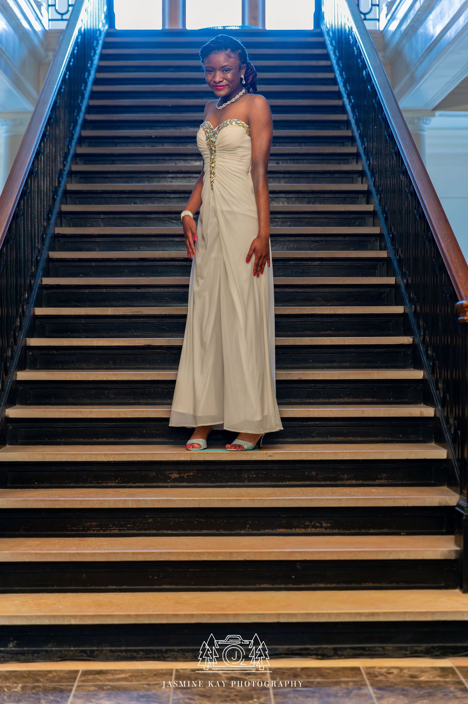 Someone in a cream-colored dress descends a grand wooden staircase in an elegant interior setting.