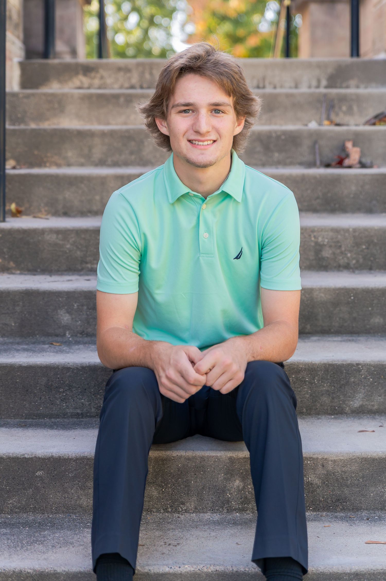 Young person in mint green polo shirt and dark pants sits casually on concrete steps outdoors.