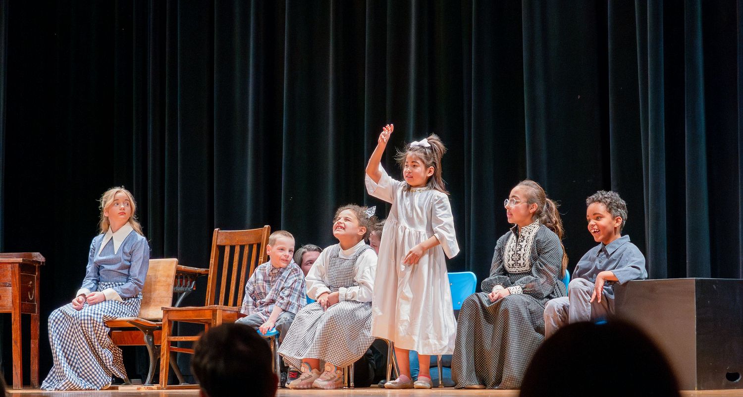 Theater performers arranged across stage in casual attire during dramatic scene.