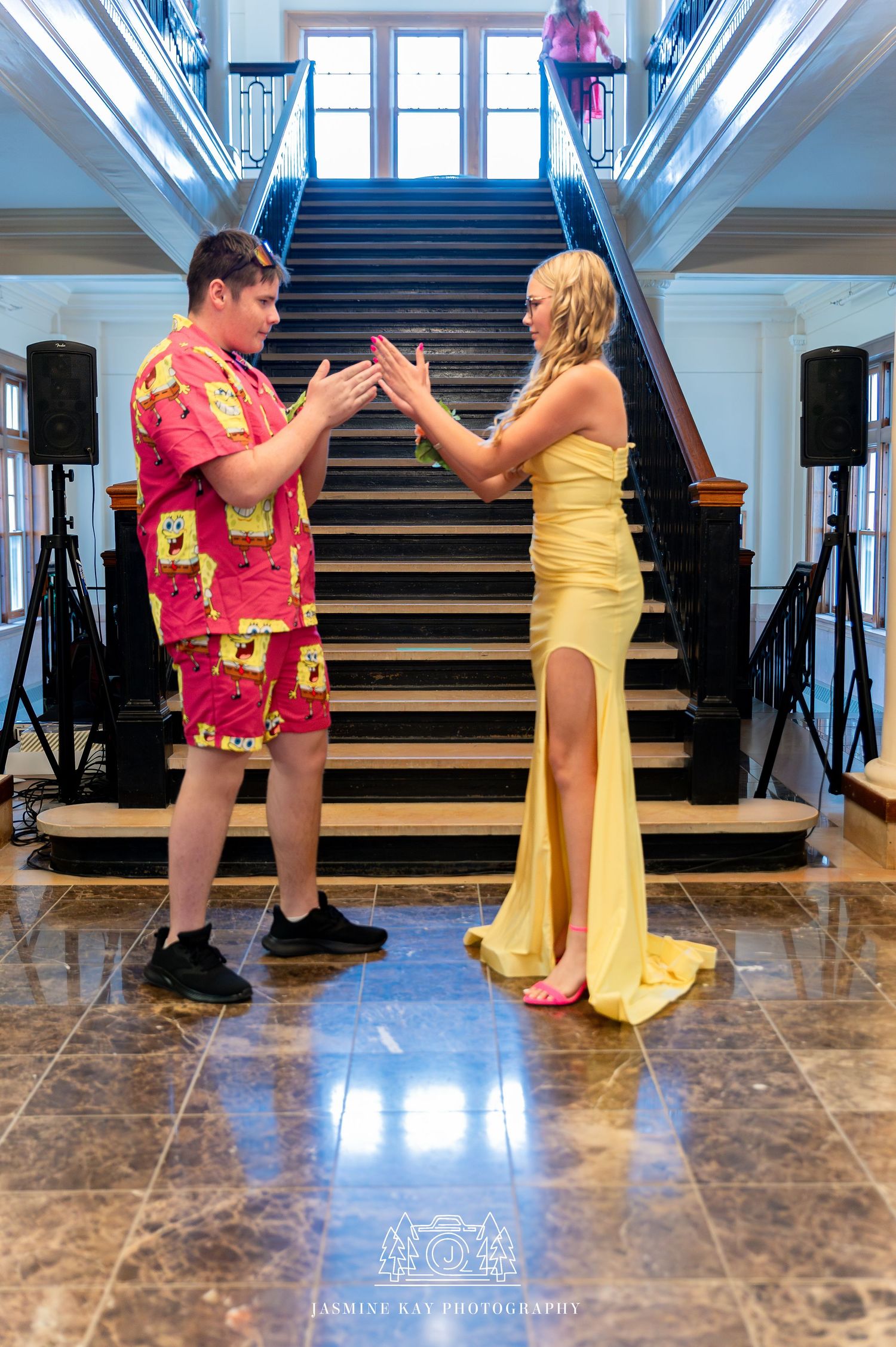 A couple in contrasting outfits - a floral Hawaiian shirt and shorts with a flowing yellow evening gown - pose on marble stairs.