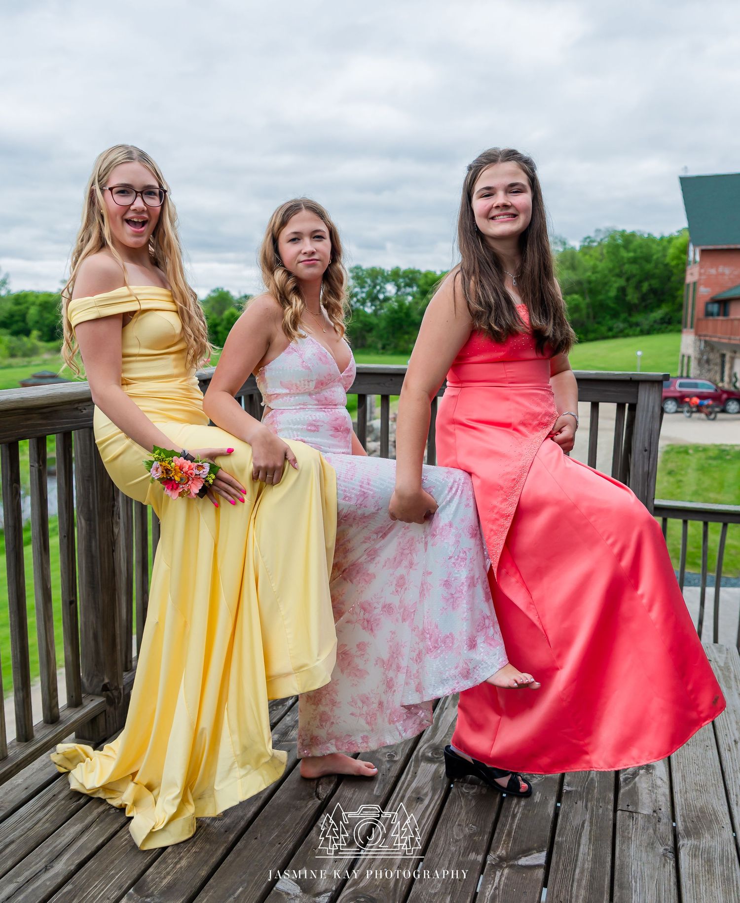 A group in formal dresses poses on a wooden deck, wearing yellow and coral colored gowns against a green landscape.