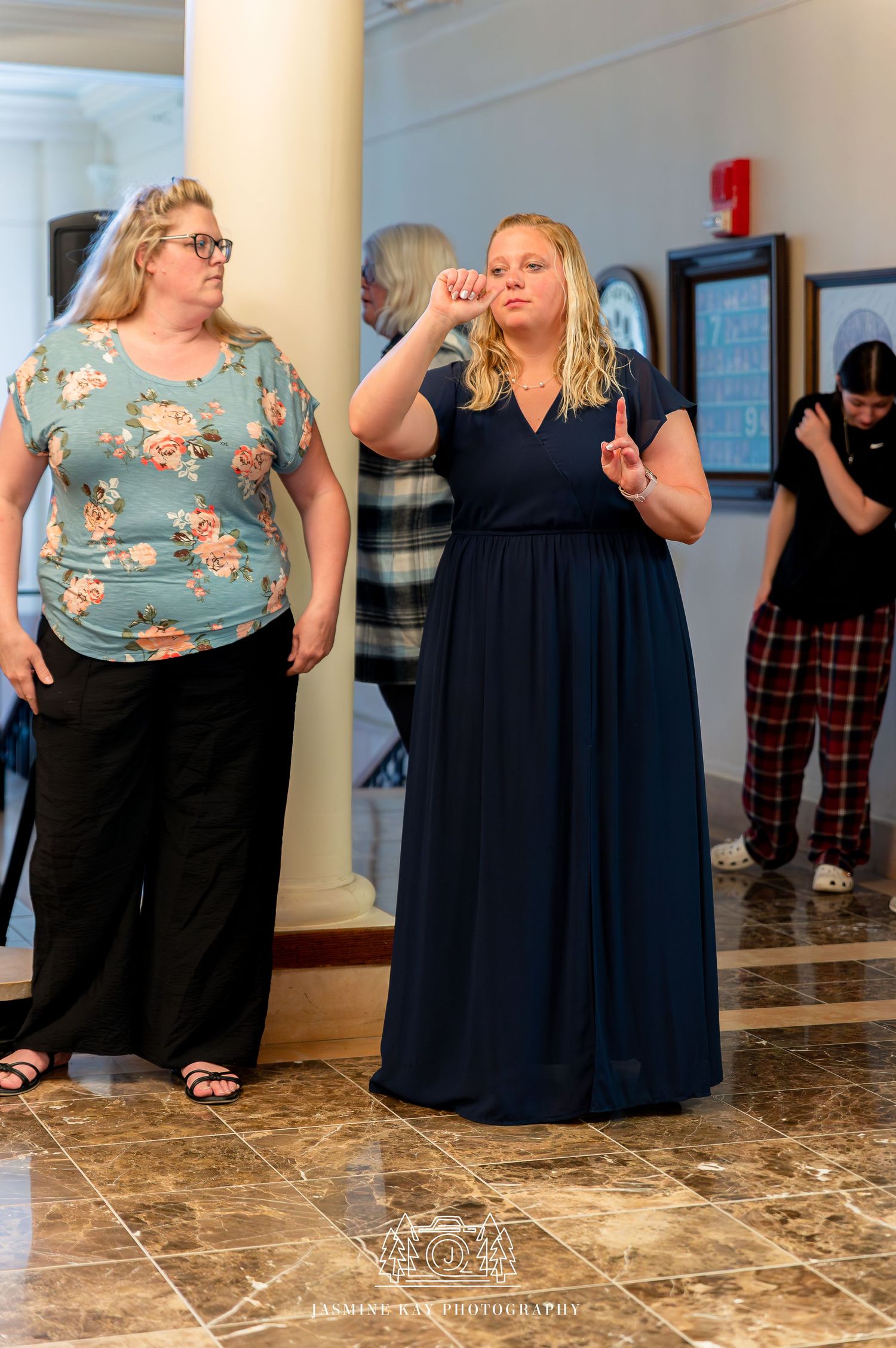 Two women in long dresses stand together in a marble-floored room, one gesturing animatedly while the other watches.
