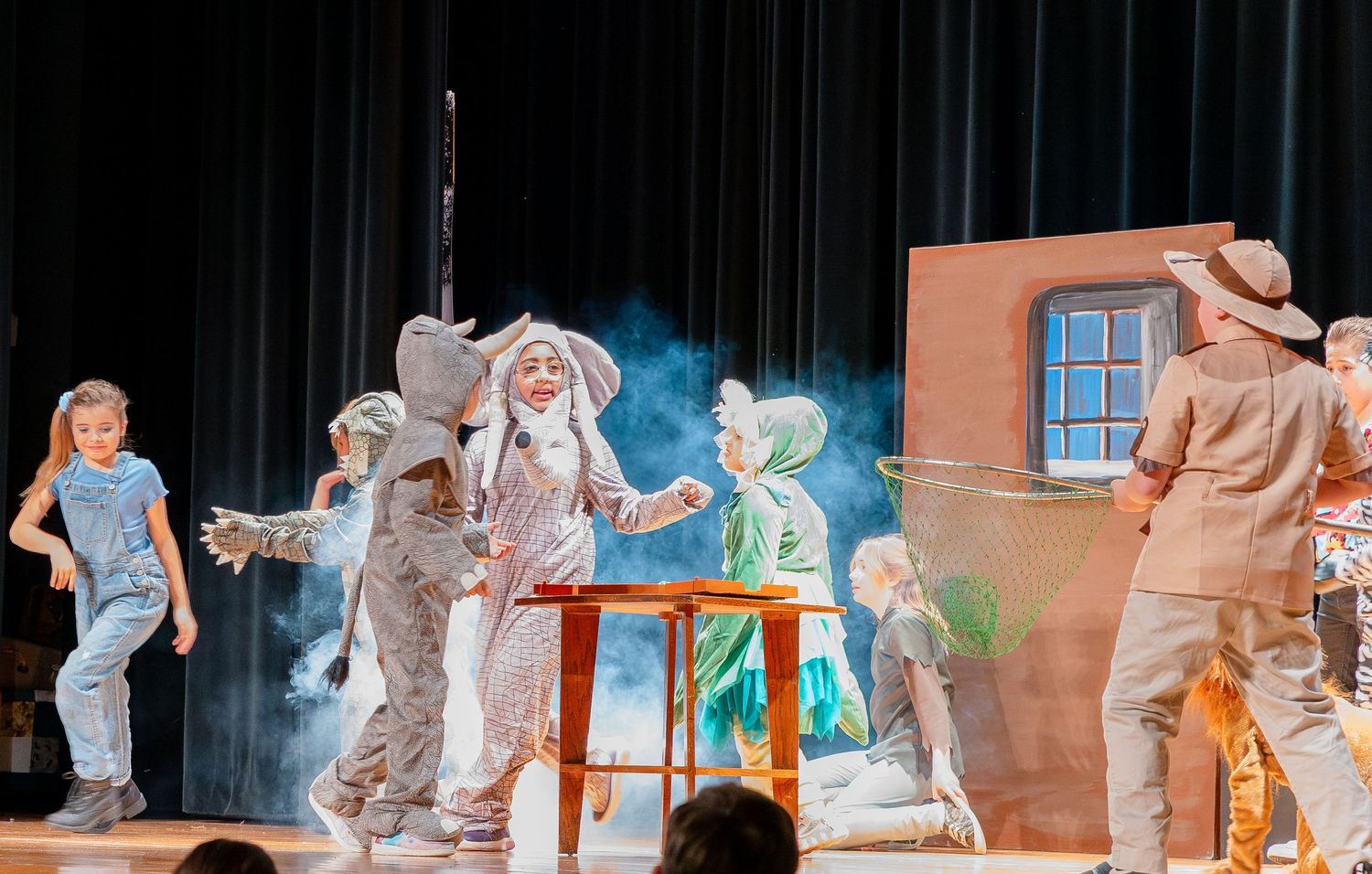 Performers act out a scene on stage with wooden chairs and benches during a theatrical production.