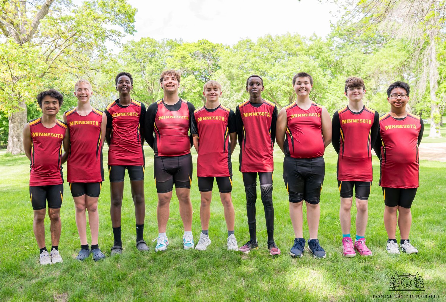 A panoramic photo of a rowing team wearing red uniforms standing in a line outdoors against a green background.