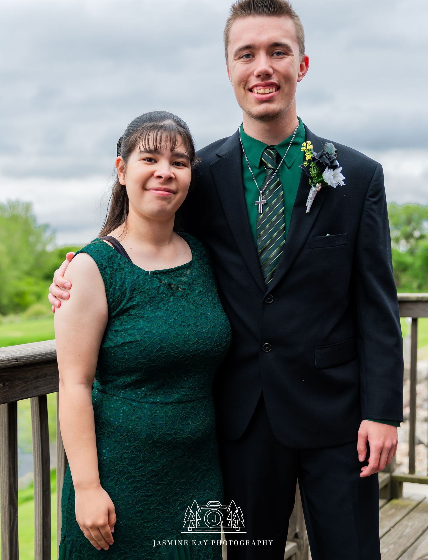 A couple in formal attire, with the woman wearing a dark green lace dress and the man in a black suit with green tie.
