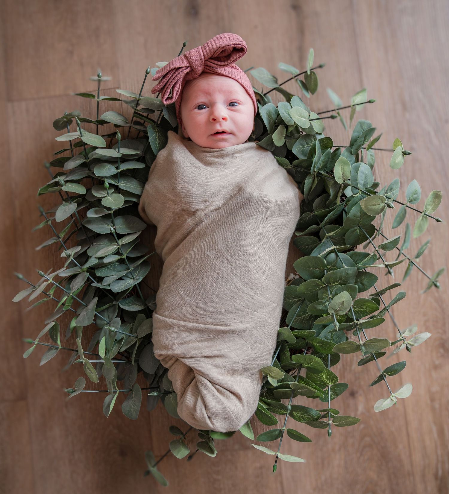 Newborn baby wrapped in cream swaddle with pink headband lying beside eucalyptus greenery on wooden floor.
