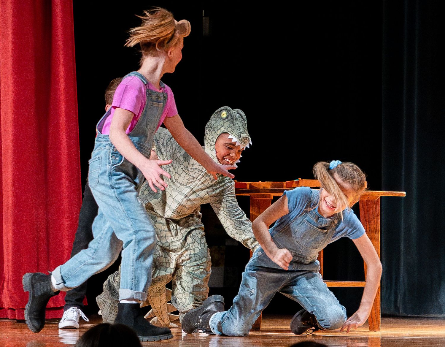 Dancers in casual attire perform energetic choreography with chairs on a stage with red curtains.