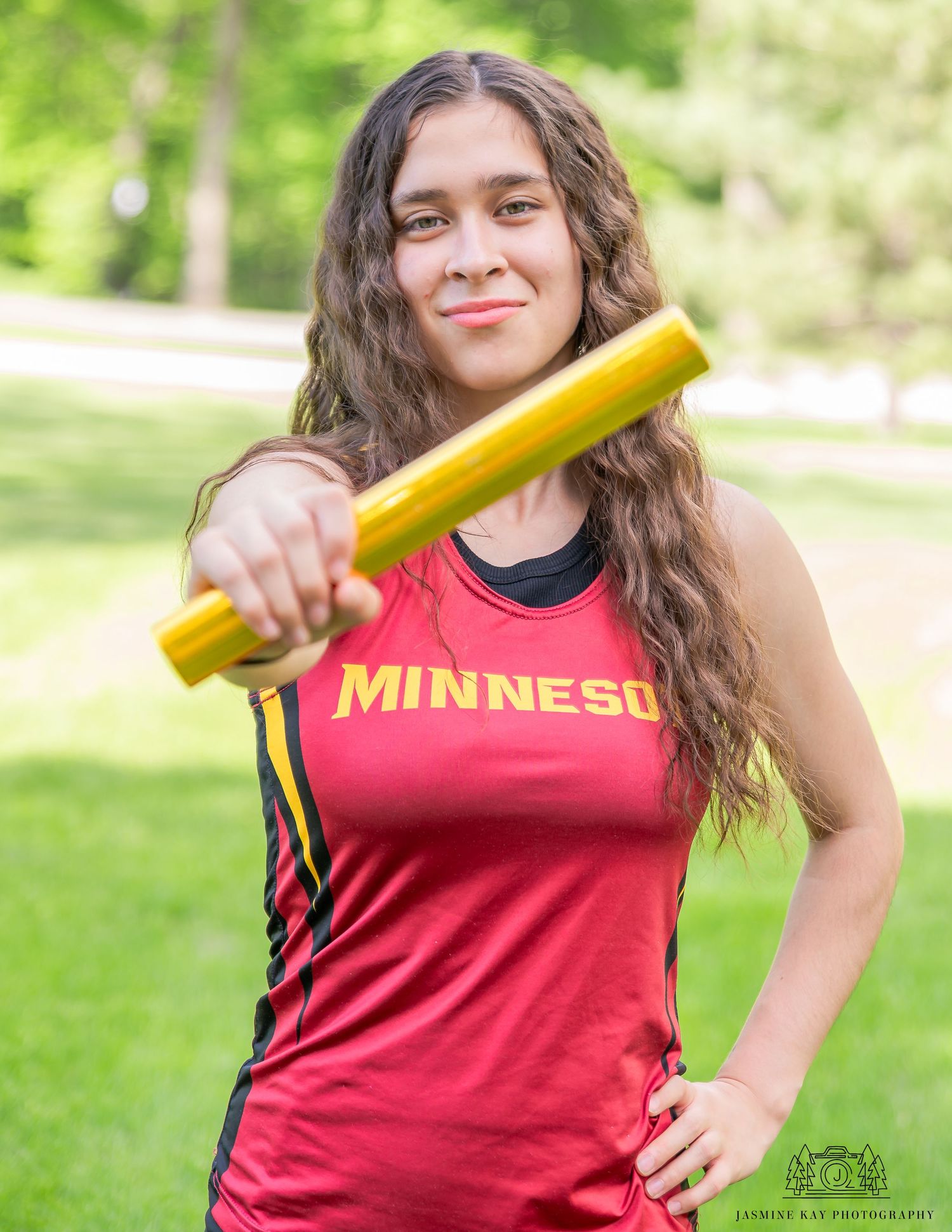 Someone in a red Minnesota shirt poses with a yellow baseball bat in a sunny outdoor setting.