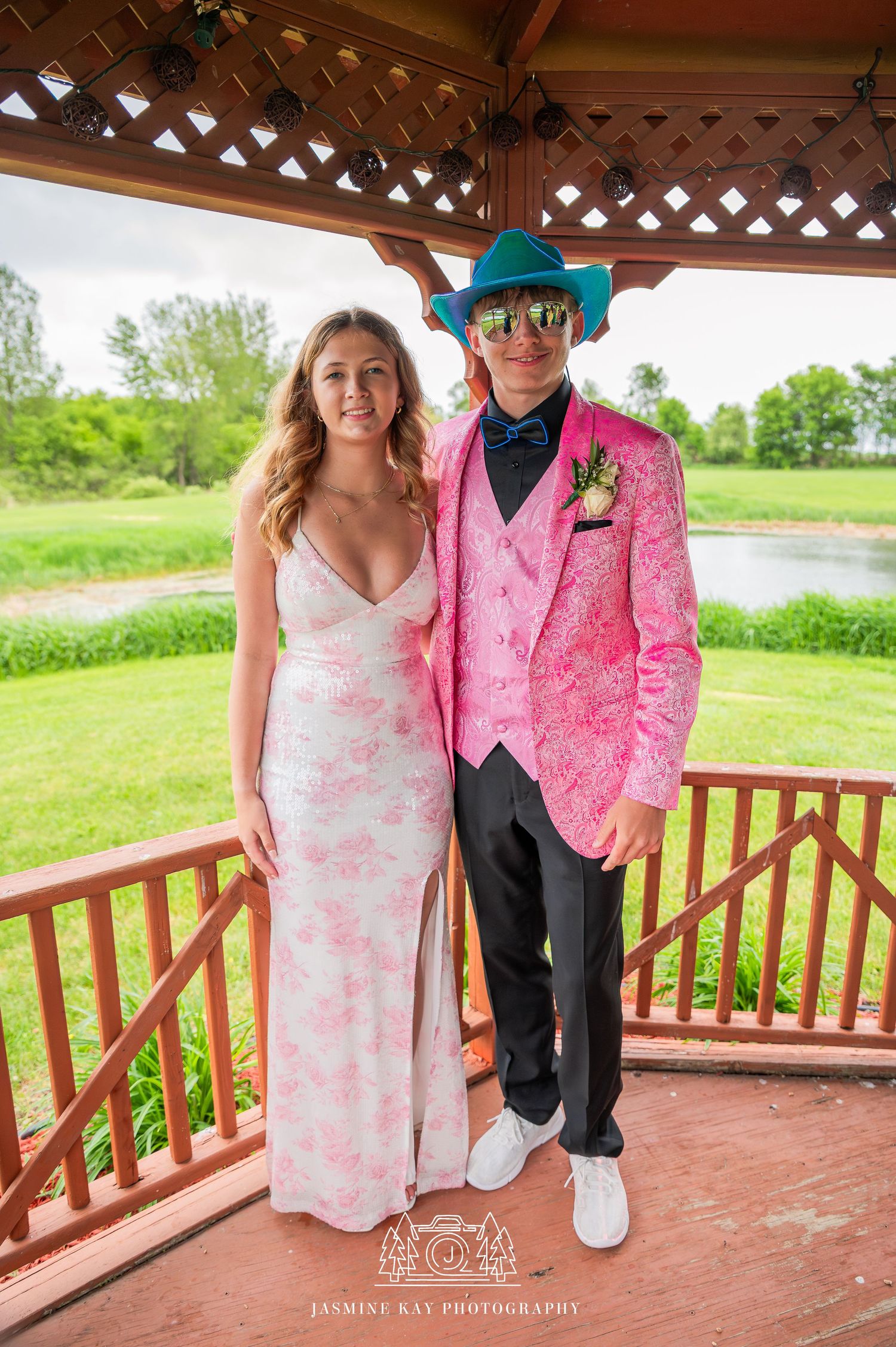 A couple in coordinating pink attire pose on a wooden gazebo deck with green scenery in the background.