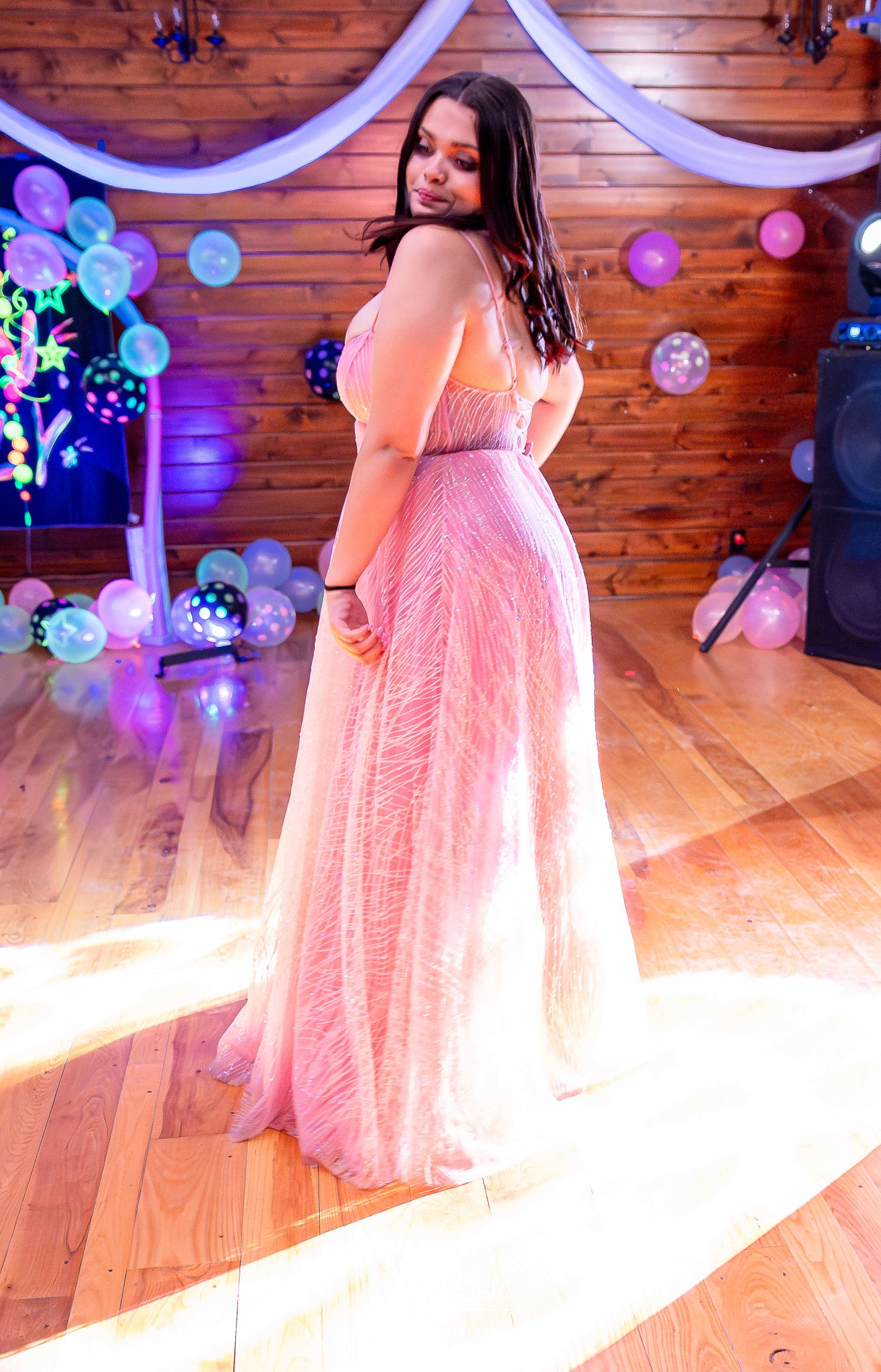 A person in a flowing pink gown twirls on a wooden dance floor decorated with balloons and purple lighting.