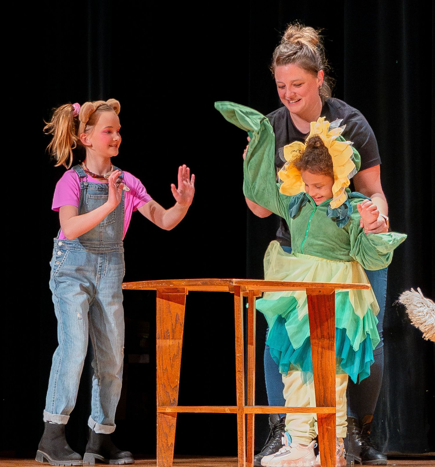 Theater scene with performers interacting around a wooden table with colorful costumes and props.