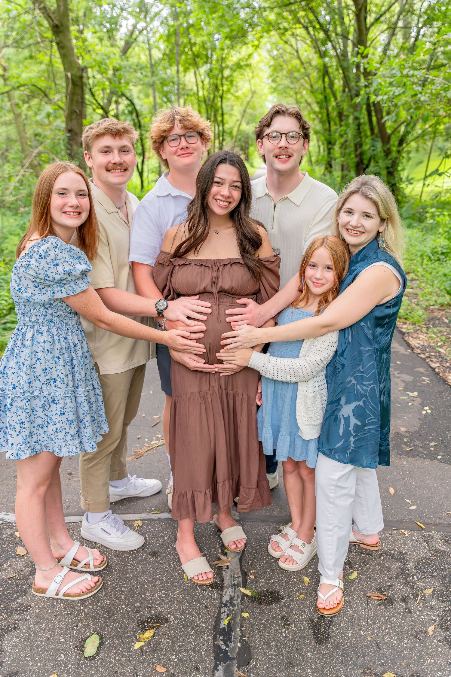 A group of people wearing casual summer outfits stand together on a wooded path in dappled sunlight.