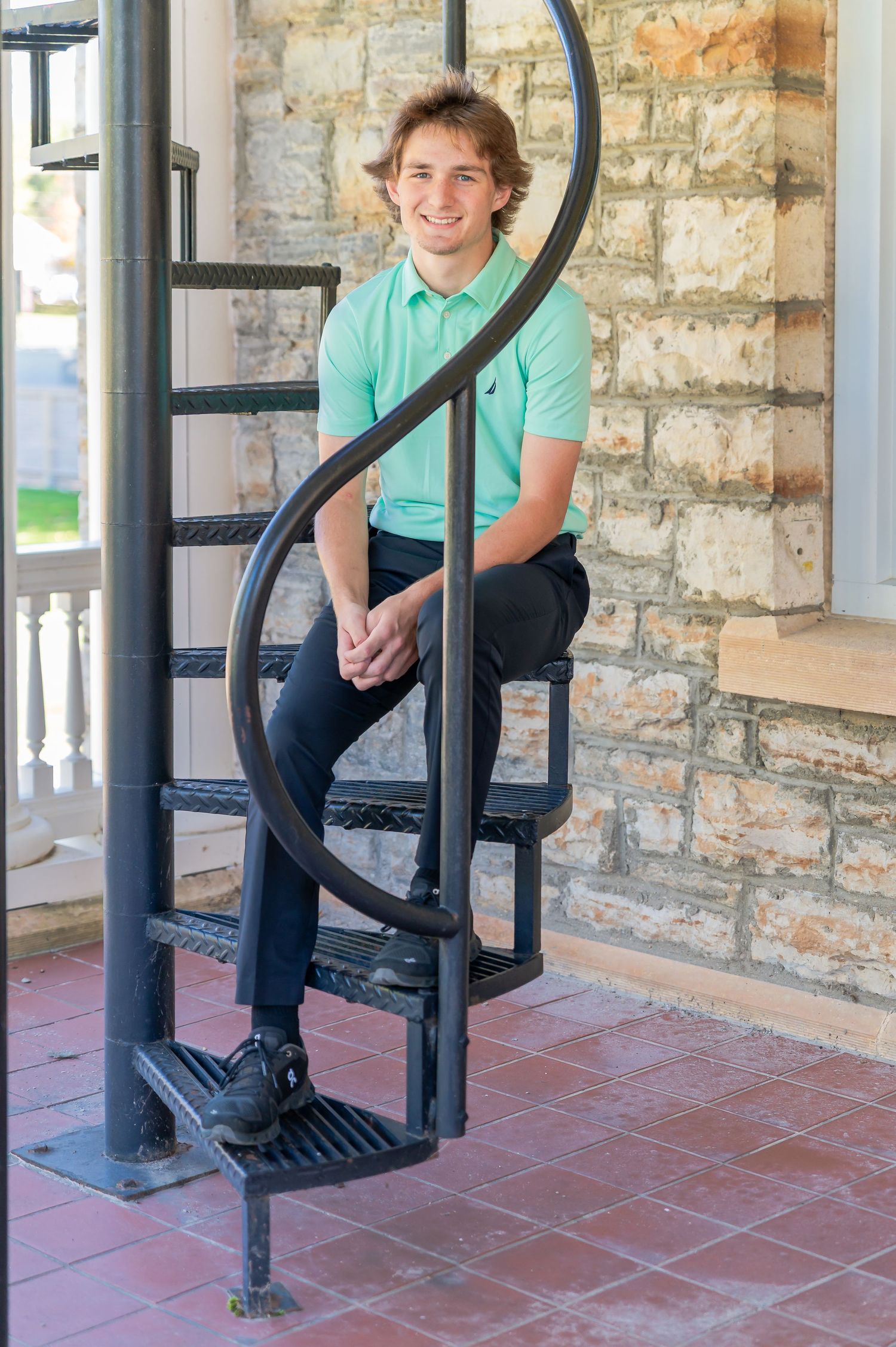 Person in casual attire poses along a stone wall with black iron railings on a tiled porch.