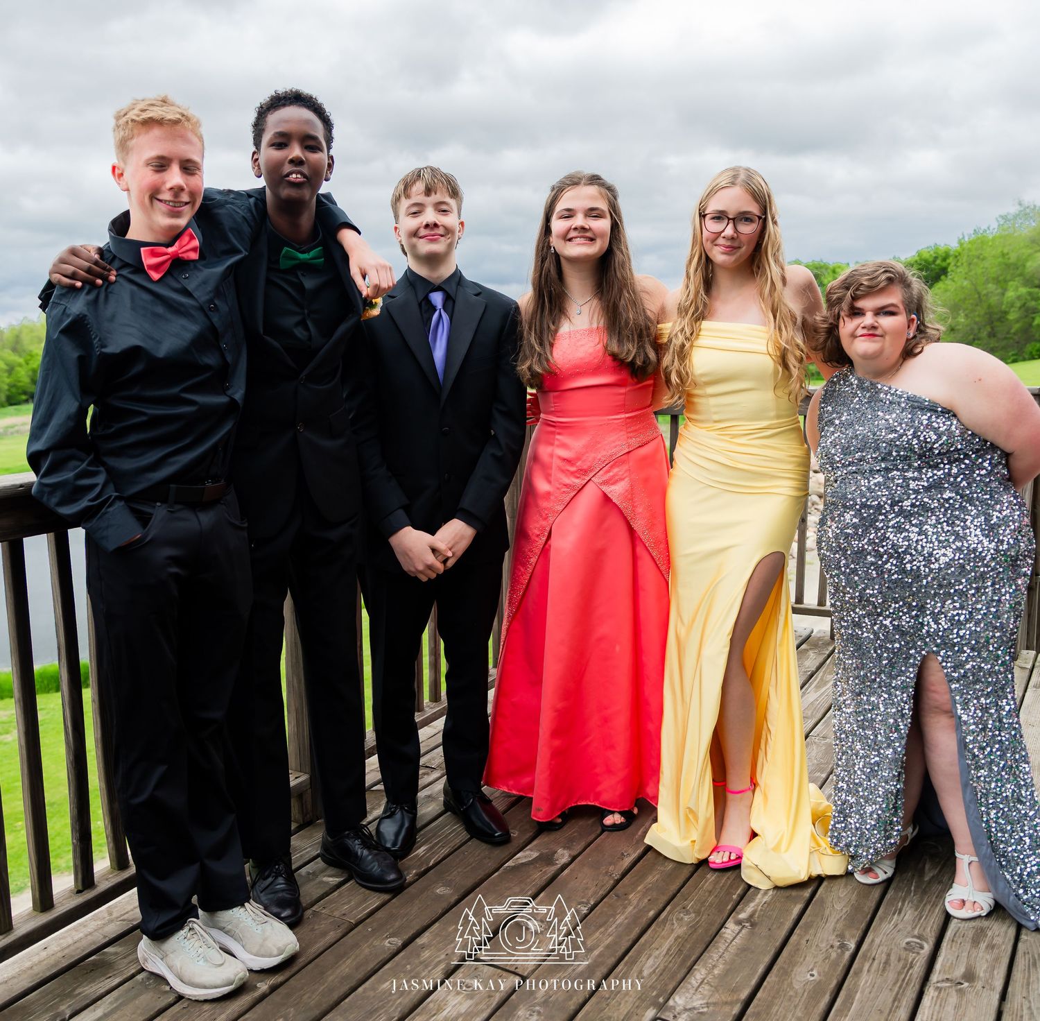 A group in formal attire including dresses and suits poses together on a wooden deck during a special occasion.
