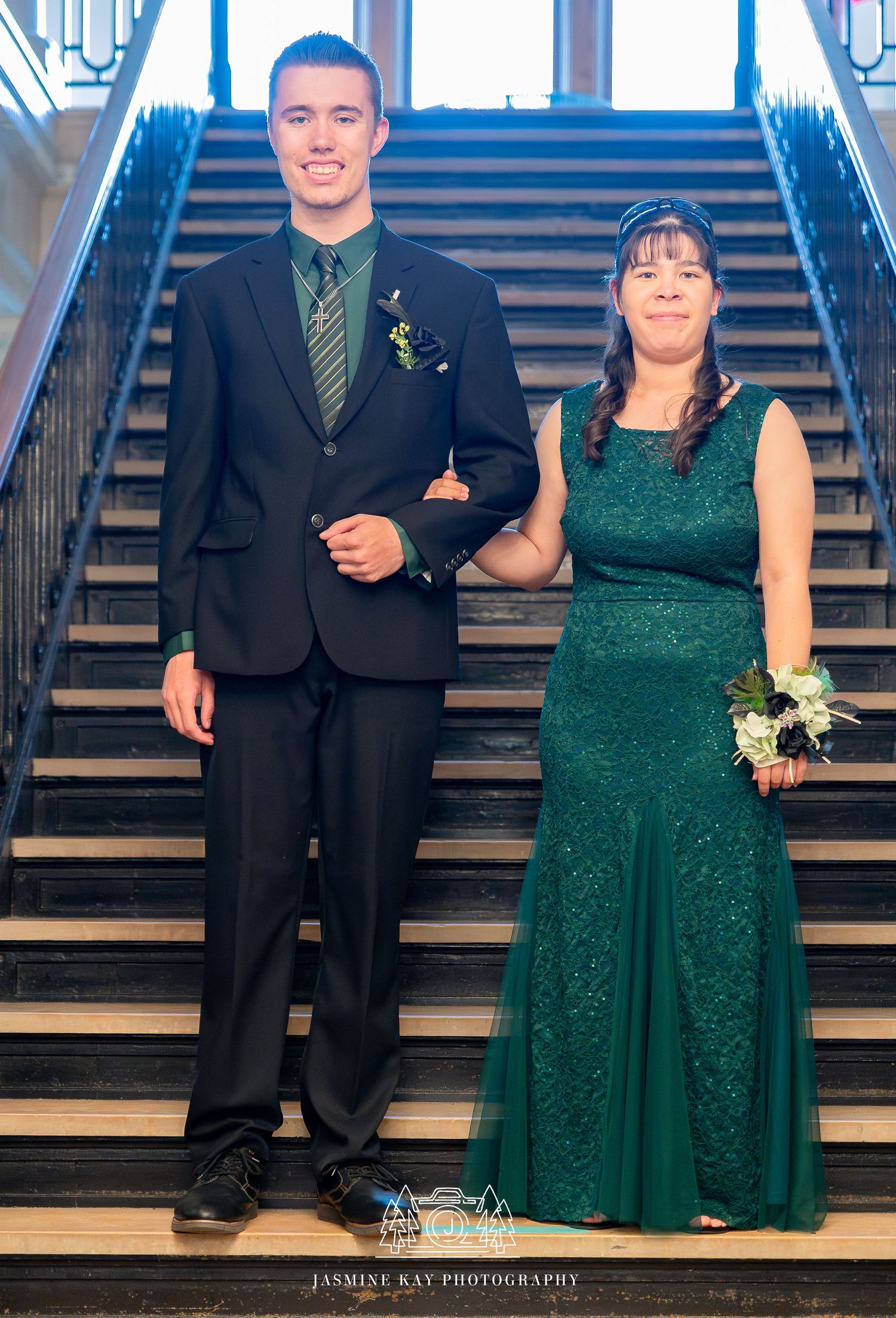 A couple poses on elegant stairs for a formal event, with the woman in emerald green and the man in a black suit.
