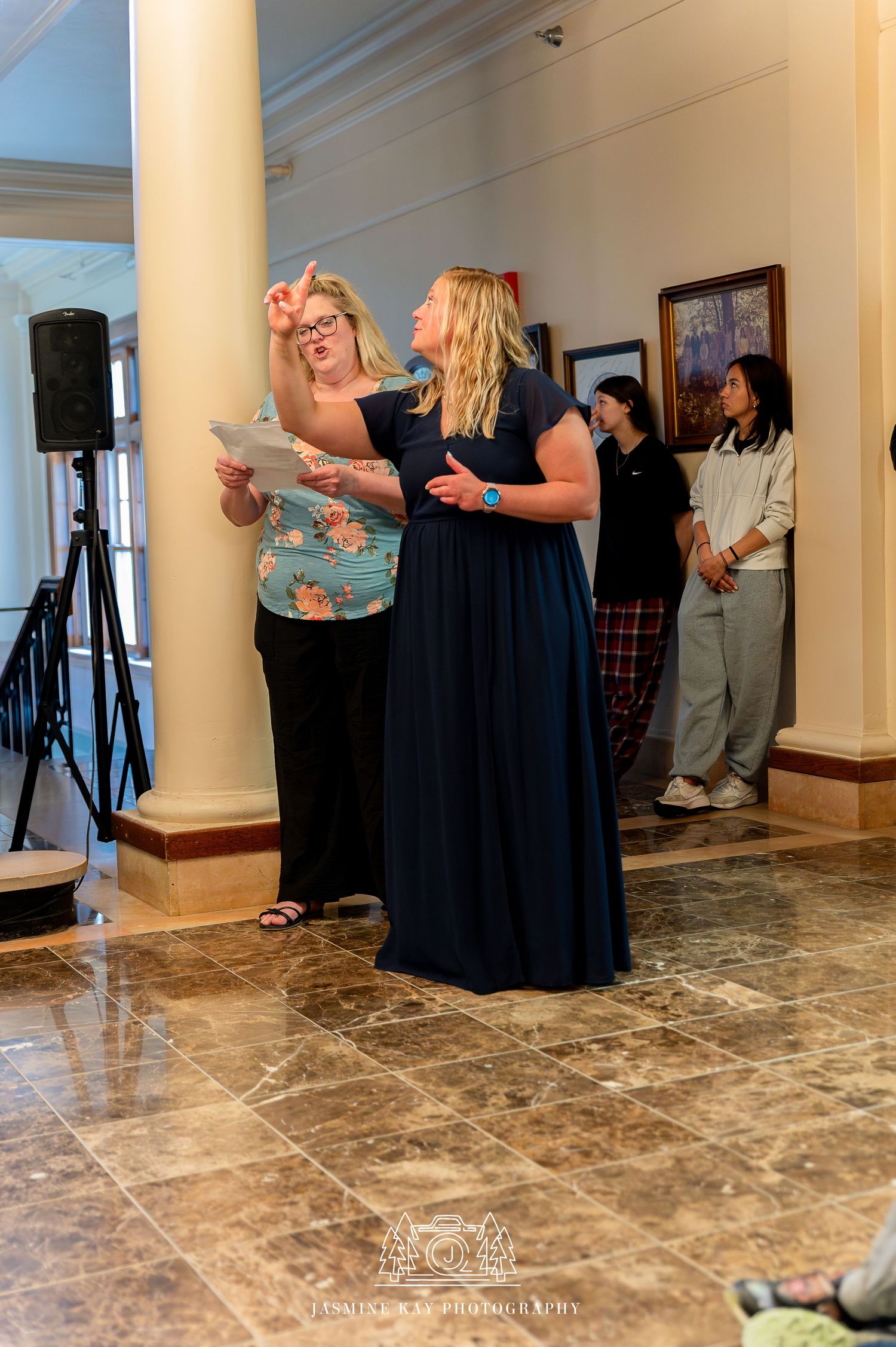 Two people stand reading from papers during what appears to be a performance or presentation in an elegant room with columns.